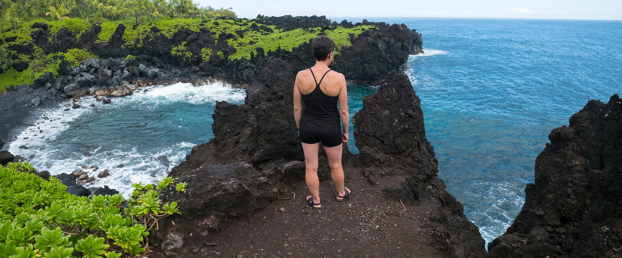 A woman standing on black volcanic rocks overlooking the ocean on a cloudy day, with lush green foliage nearby in Maui.