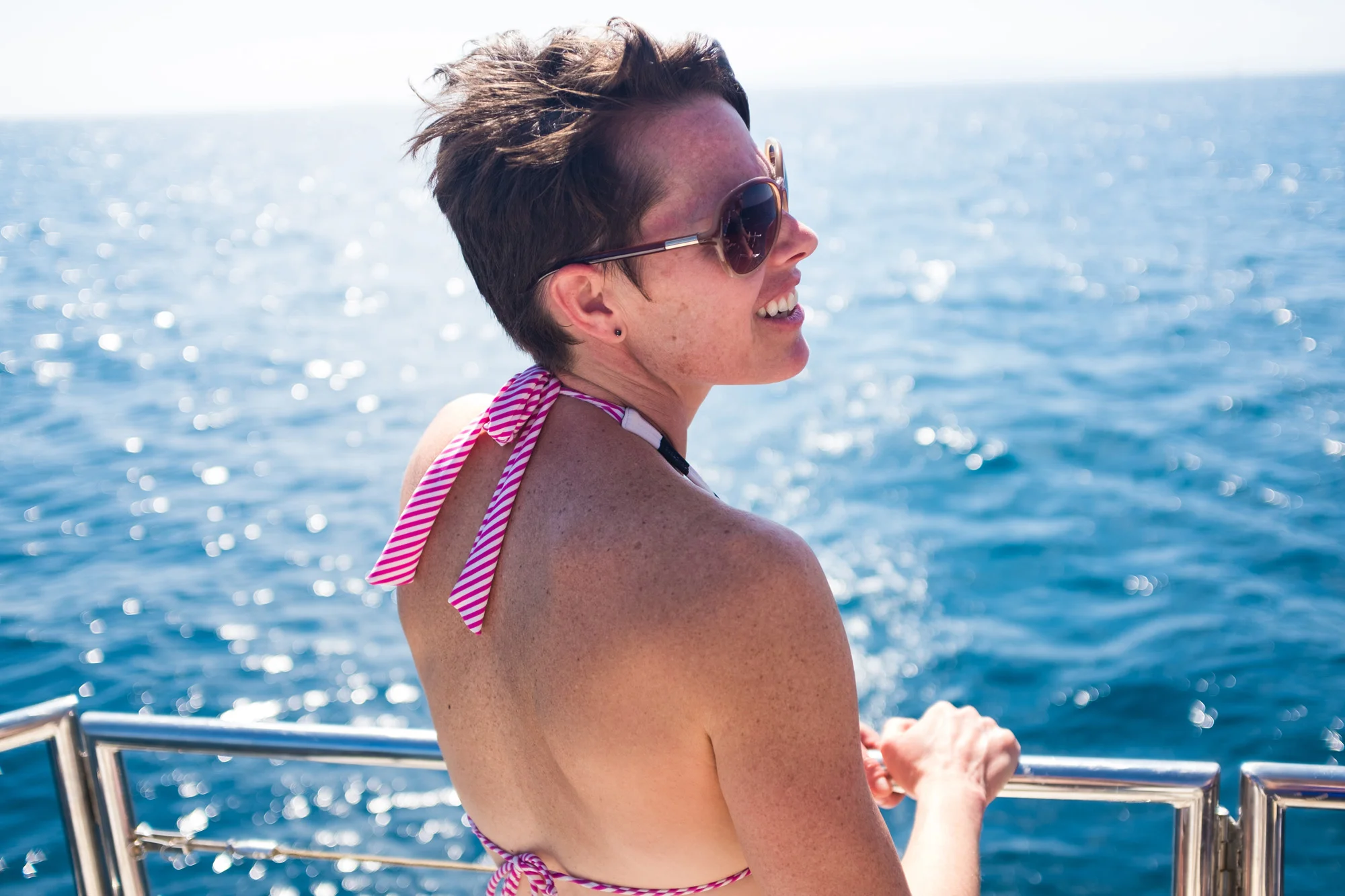A woman with short brown hair wearing sunglasses and a pink and white striped bikini, leaning on a boat railing with the ocean in the background  in Maui.