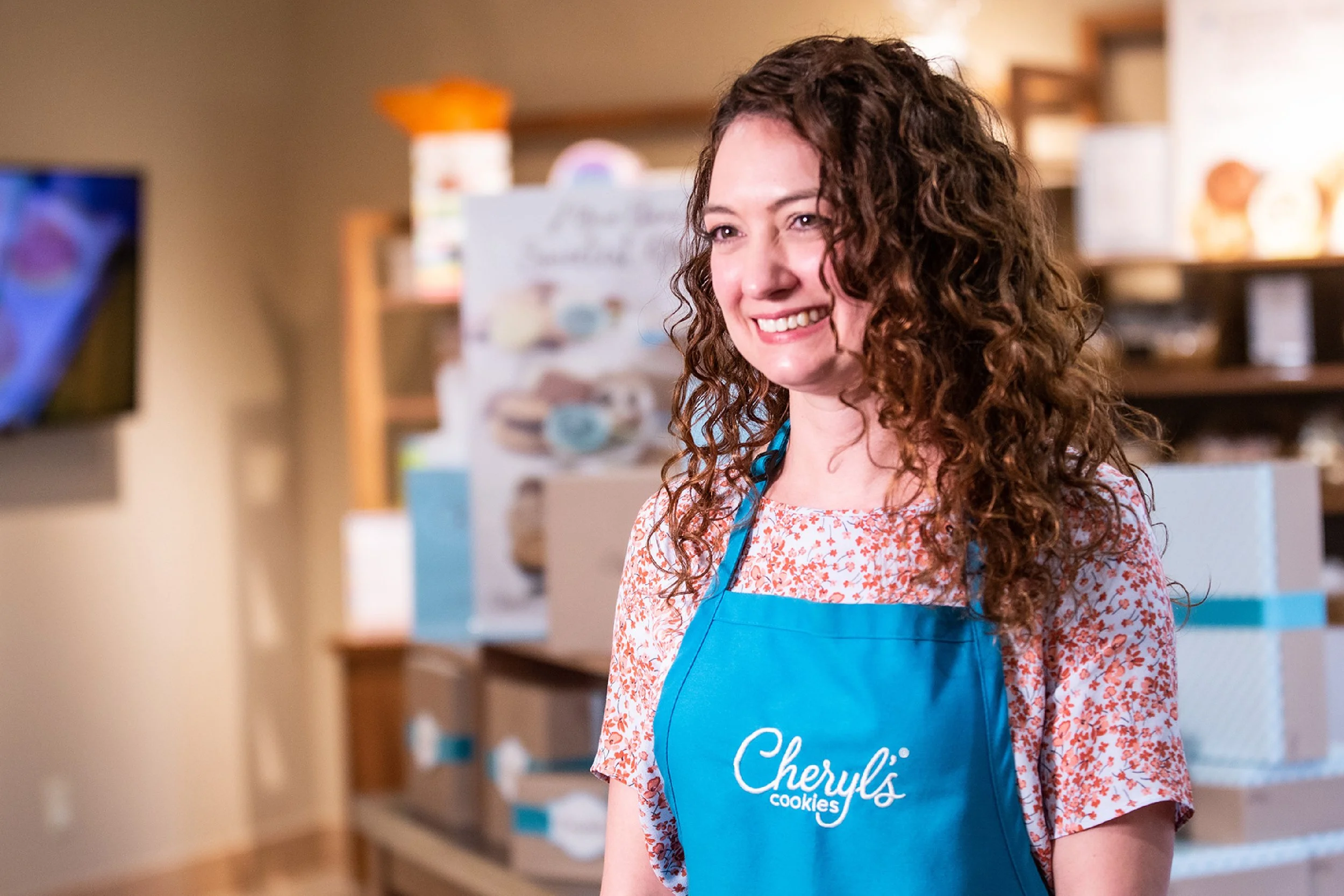 A woman with curly brown hair wearing a blue apron with 'Cheryl's Cookies' written on it, smiling in a bakery or cookie shop.
