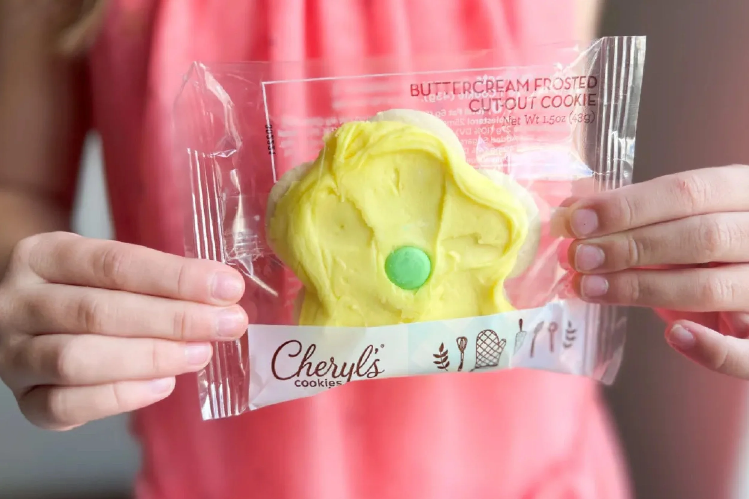 Person holding a packaged yellow buttercream frosted cut-out cookie with a green dot on it, labeled 'Cheryl's cookies'.