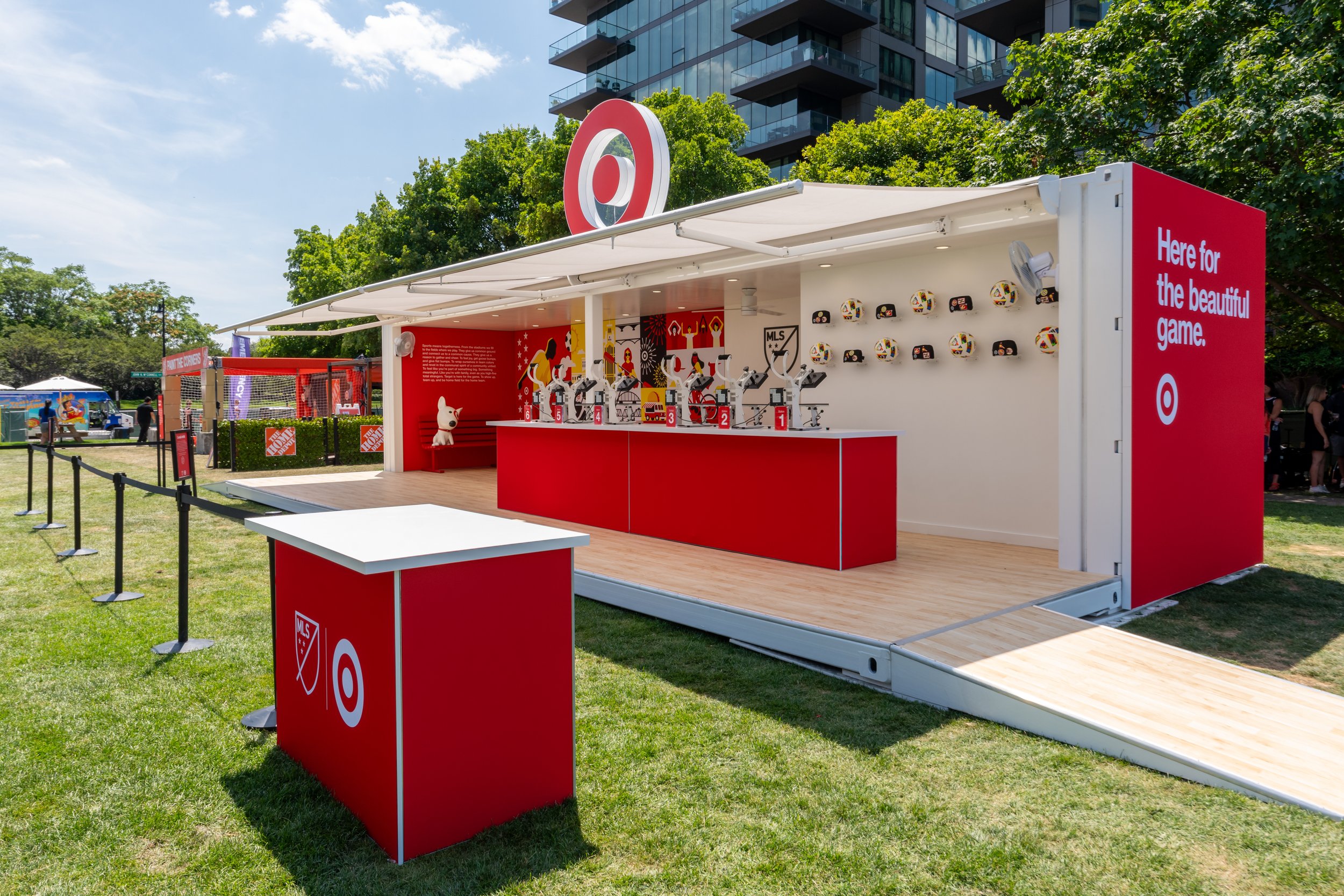 A white mobile exhibit with a red interior, displaying Target and MLS branding, set up outdoors with green grass and trees around. The display features stationary bikes, soccer helmets, and a cartoon dog mascot. There's a ramp leading into the exhibi