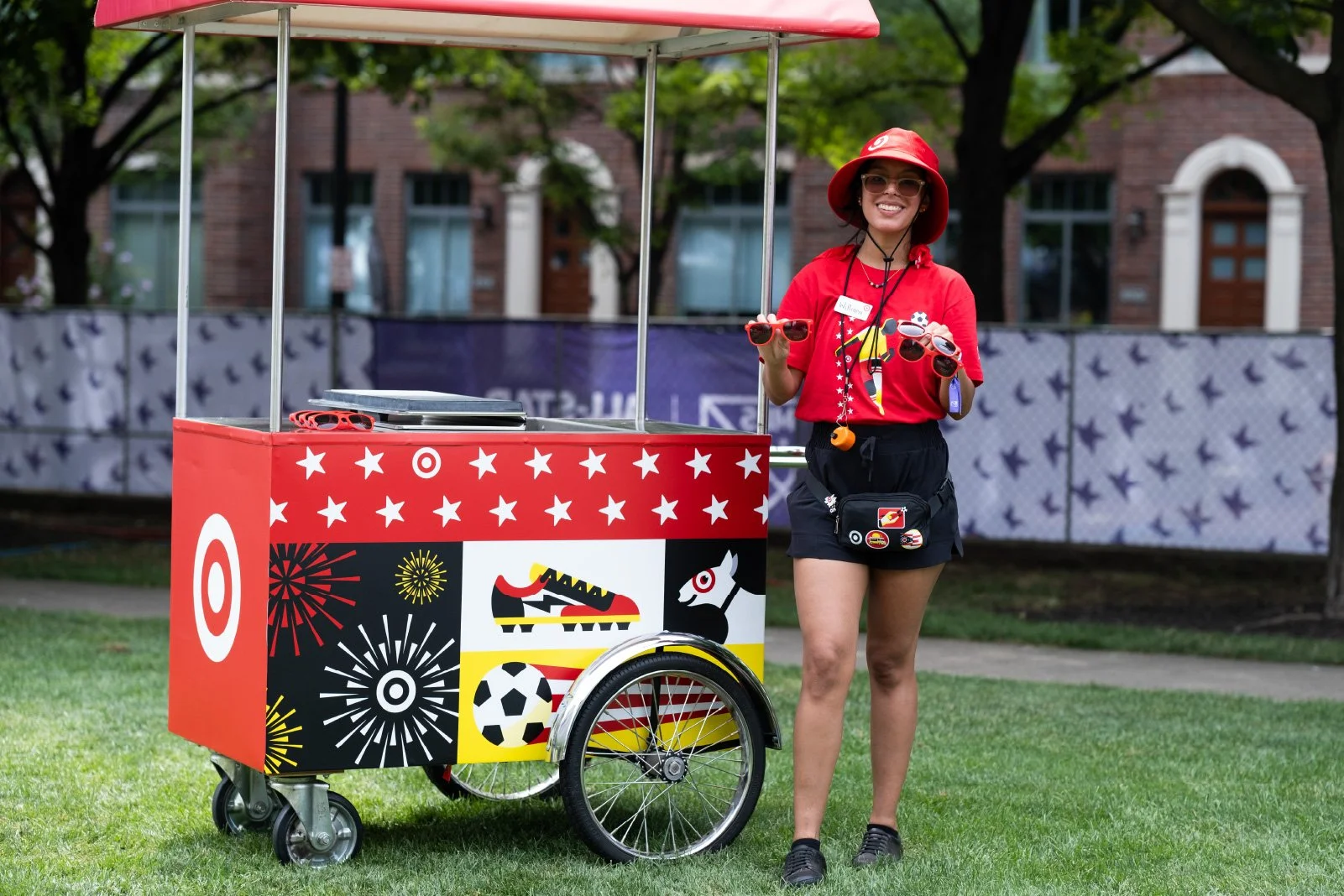 A young woman dressed in red and black, wearing a red hat and sunglasses, standing next to a colorful target-themed ice cream cart on a grassy area outdoors. She is holding a pair of sunglasses and smiling.