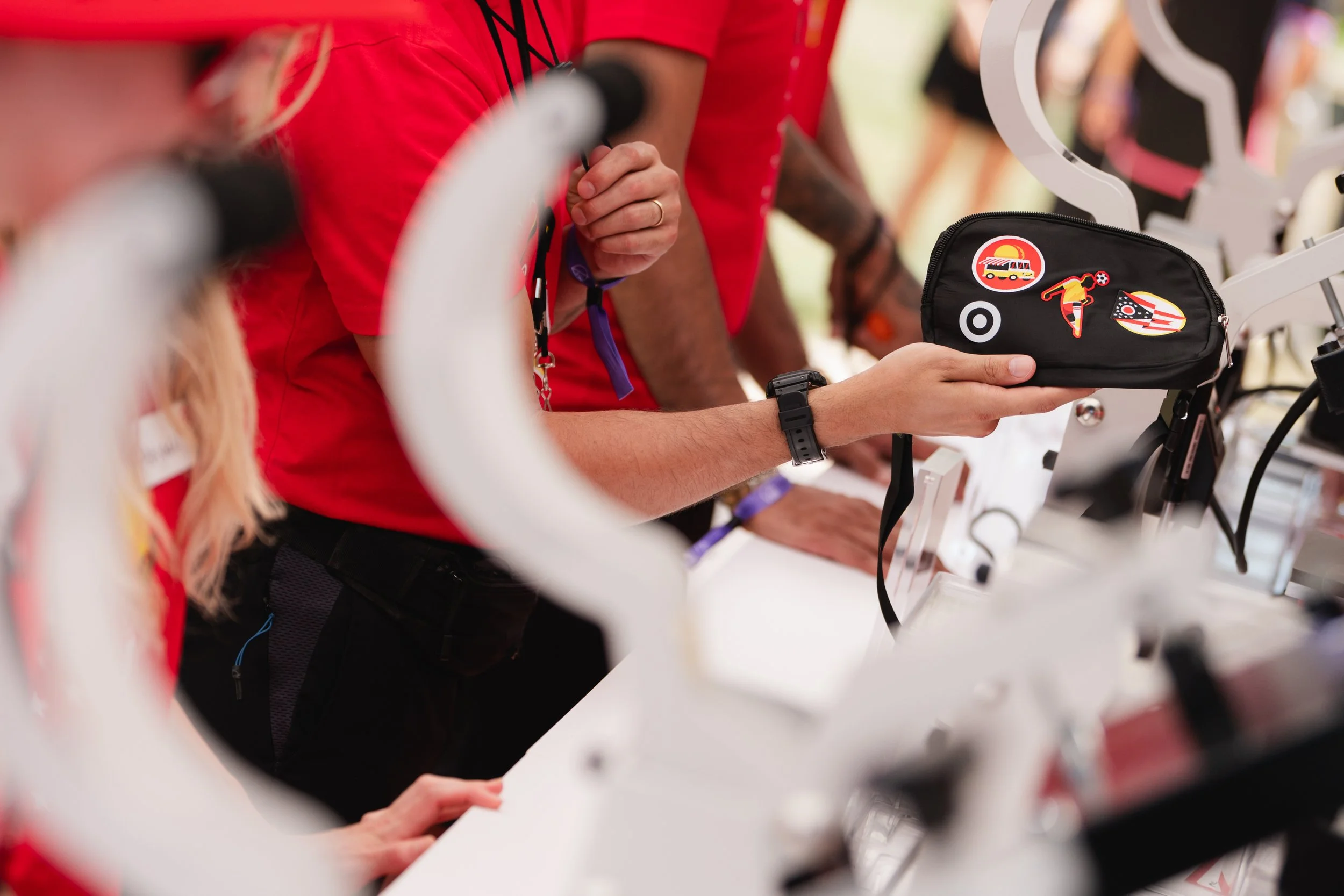 People at a booth examining merchandise, with one person holding a black zippered pouch decorated with patches, including a bus, a skateboard, and stars, with bicycle handlebars in the foreground.