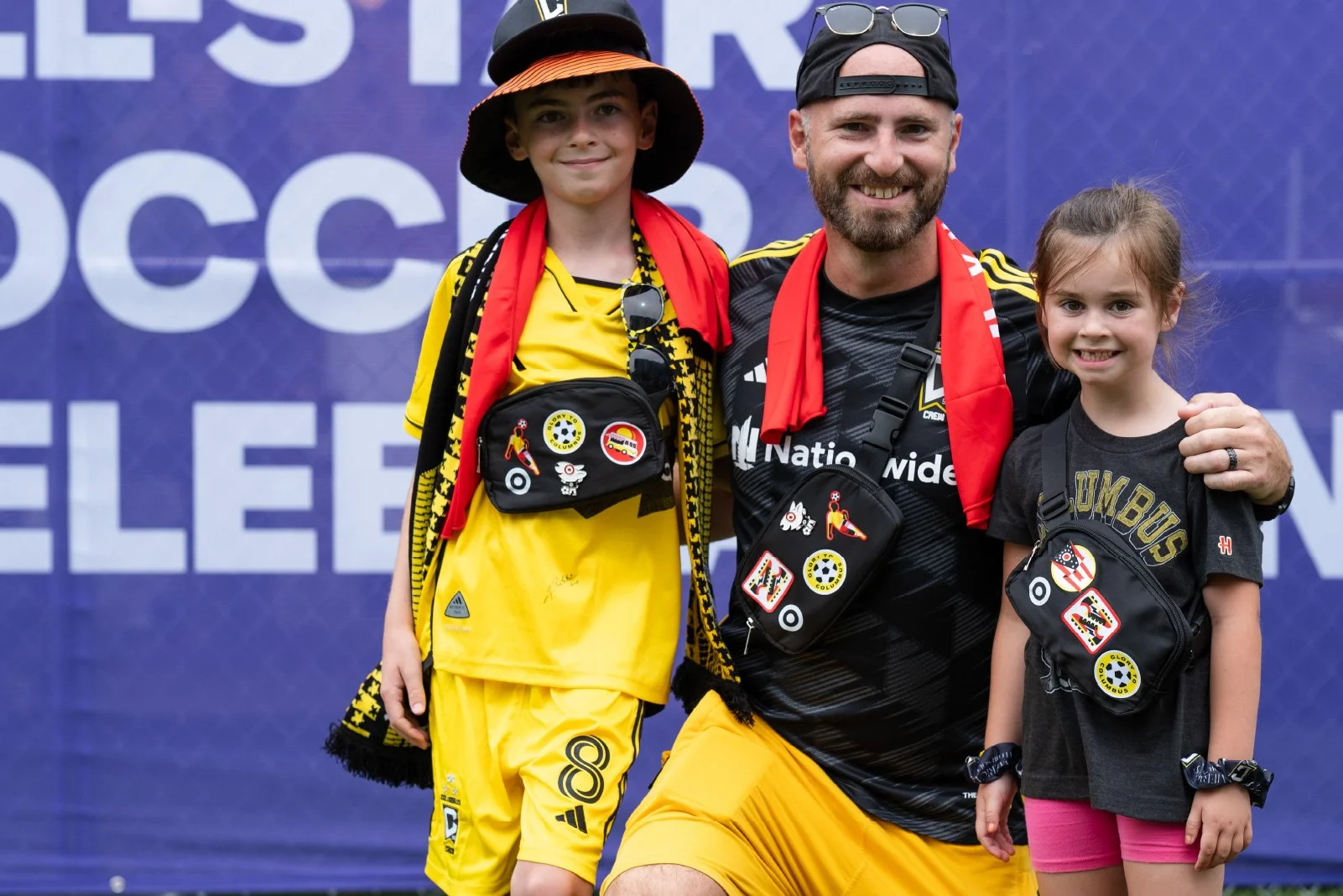 A man and two children standing together at a soccer event, all wearing black and yellow soccer gear, with red scarves, black bags with patches, and smiling.
