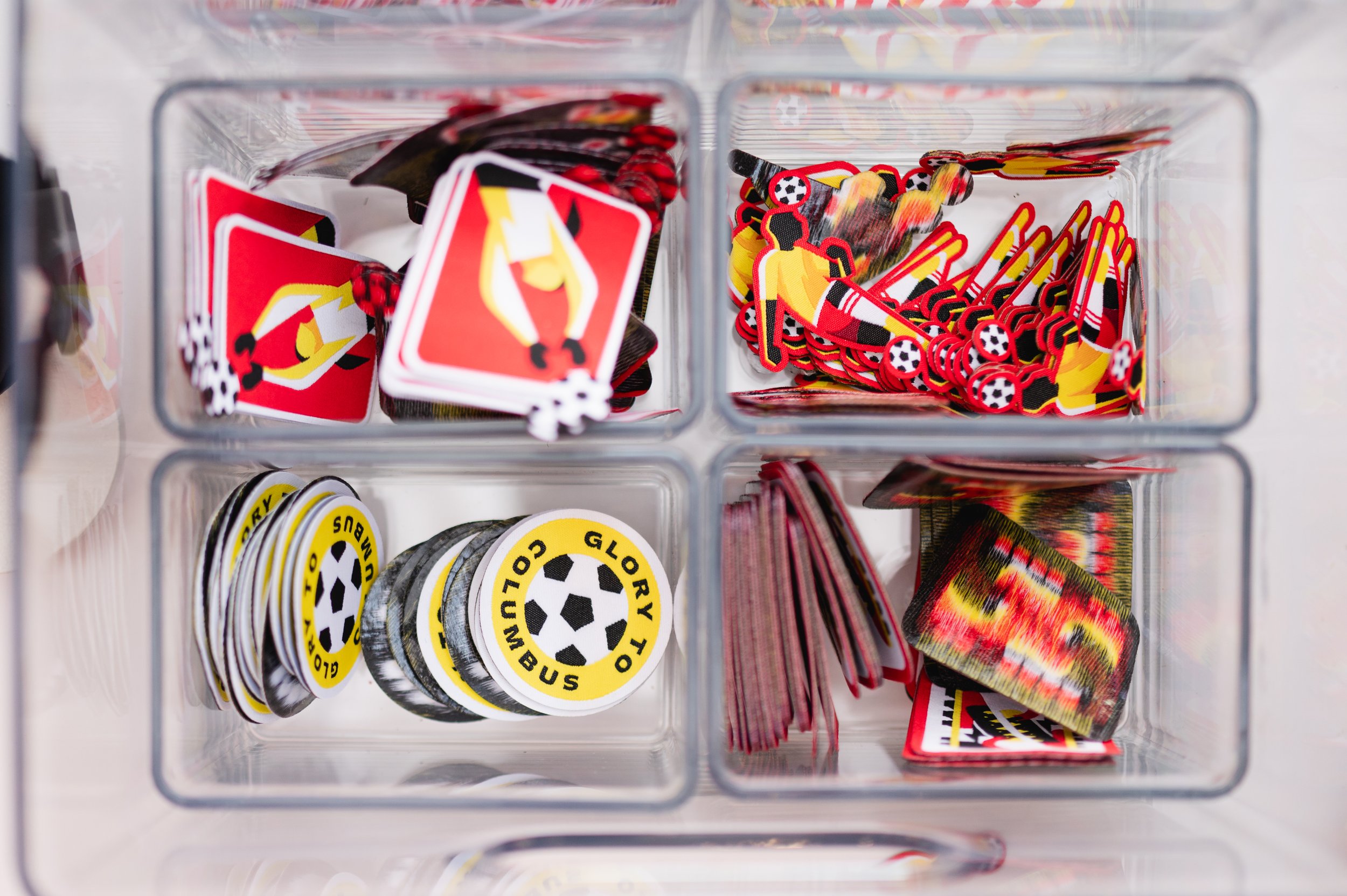 A plastic container with four sections filled with various soccer-themed game tokens and markers, including cards, circular tokens, and rectangular pieces, in red, yellow, black, and white colors.