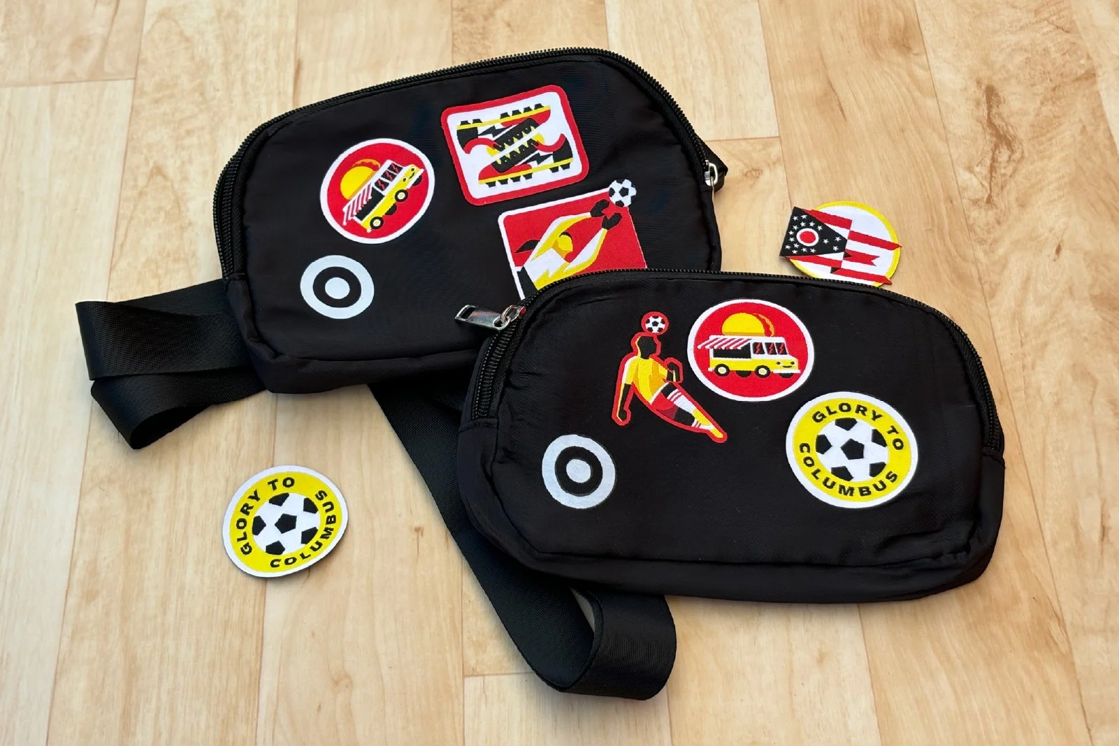 Two black bags decorated with soccer-themed patches, on a wooden floor, with additional patches and badges nearby.
