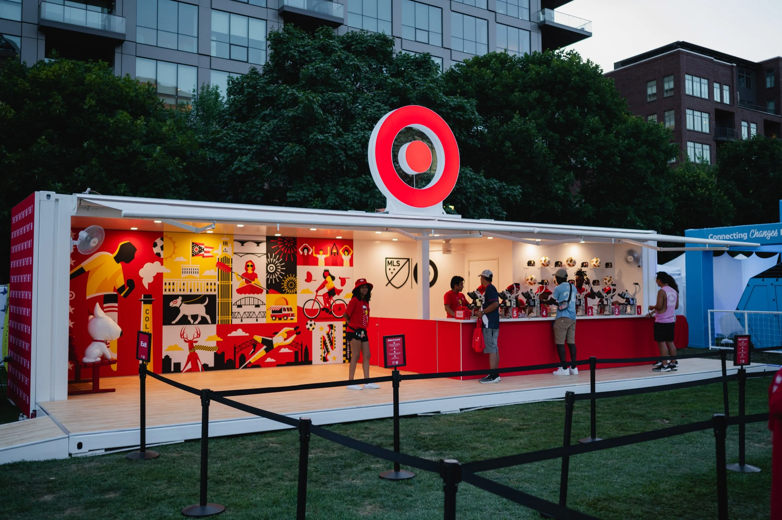A mobile event booth with a Target logo on top, featuring colorful murals of cityscapes, animals, and soccer players. Staff and visitors are interacting at a counter with themed merchandise, set outdoors near grass and trees under evening lighting.