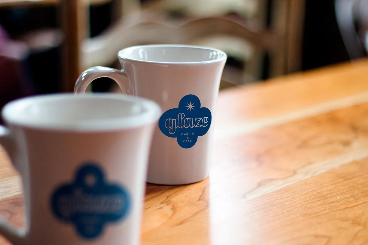 Two white ceramic mugs with blue logo of glazed bakery & cafe on a wooden table in a cafe setting, slightly out of focus.