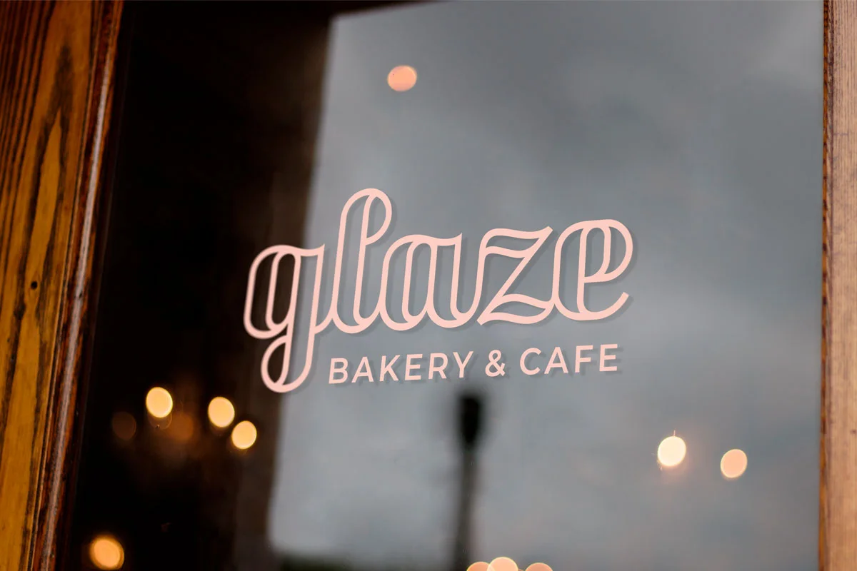 Sign for Glaze Bakery & Cafe seen through a glass window with wood framing, reflections of lights and cloudy sky outside