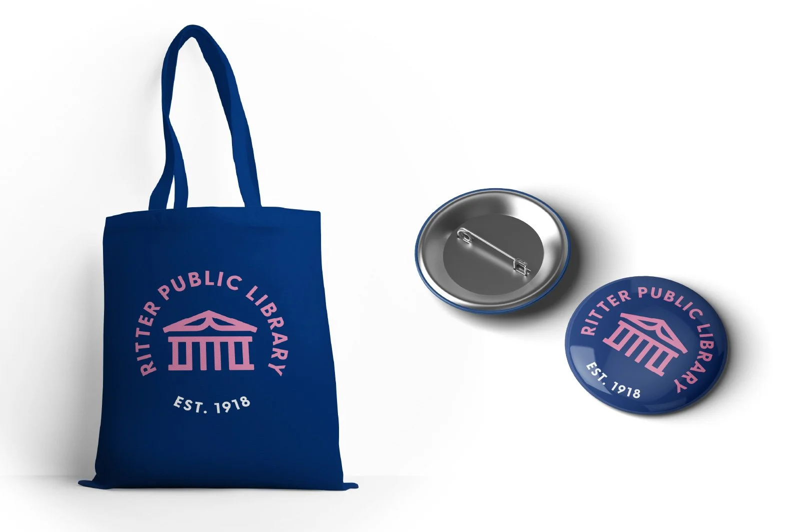 Blue tote bag with Ritter Public Library logo, and a round button with the same logo, and a pin with the logo, on a white background.