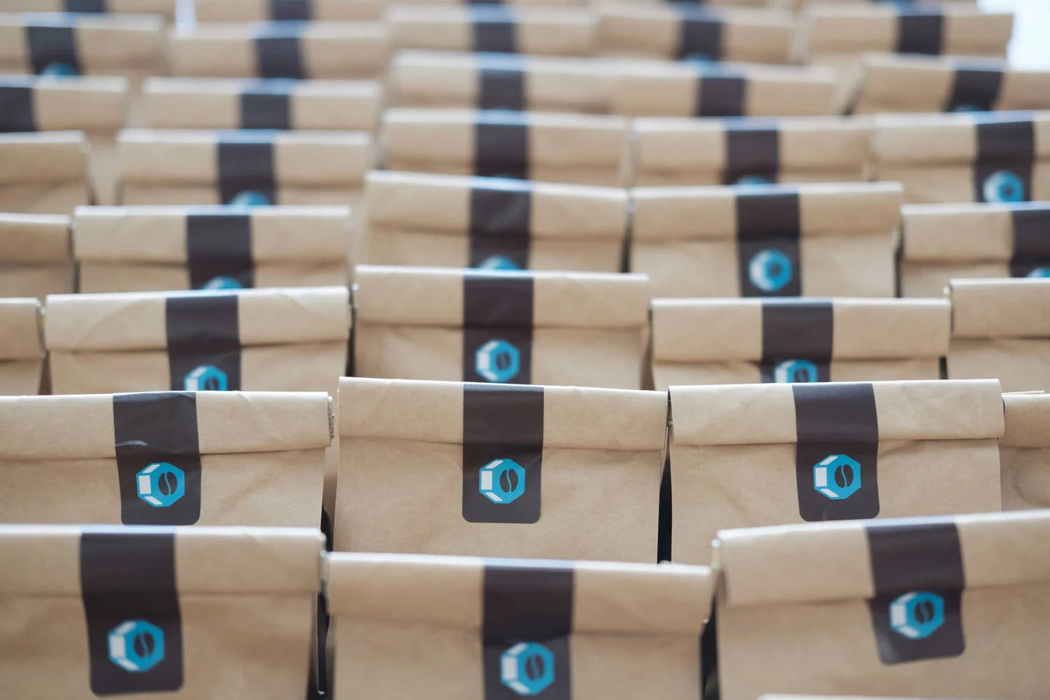 Multiple paper bags arranged in rows with a black label featuring a blue hexagon logo with a coffee bean in the center.