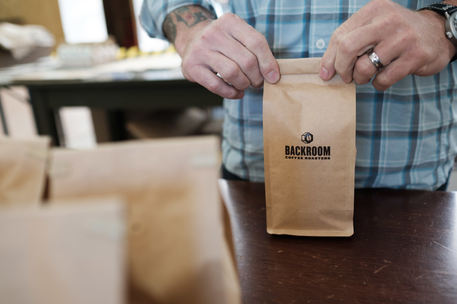A person with tattoos and a wedding ring is folding a brown paper bag with the logo and text 'Backroom Coffee Roasters' on it, in a coffee shop or roastery setting.