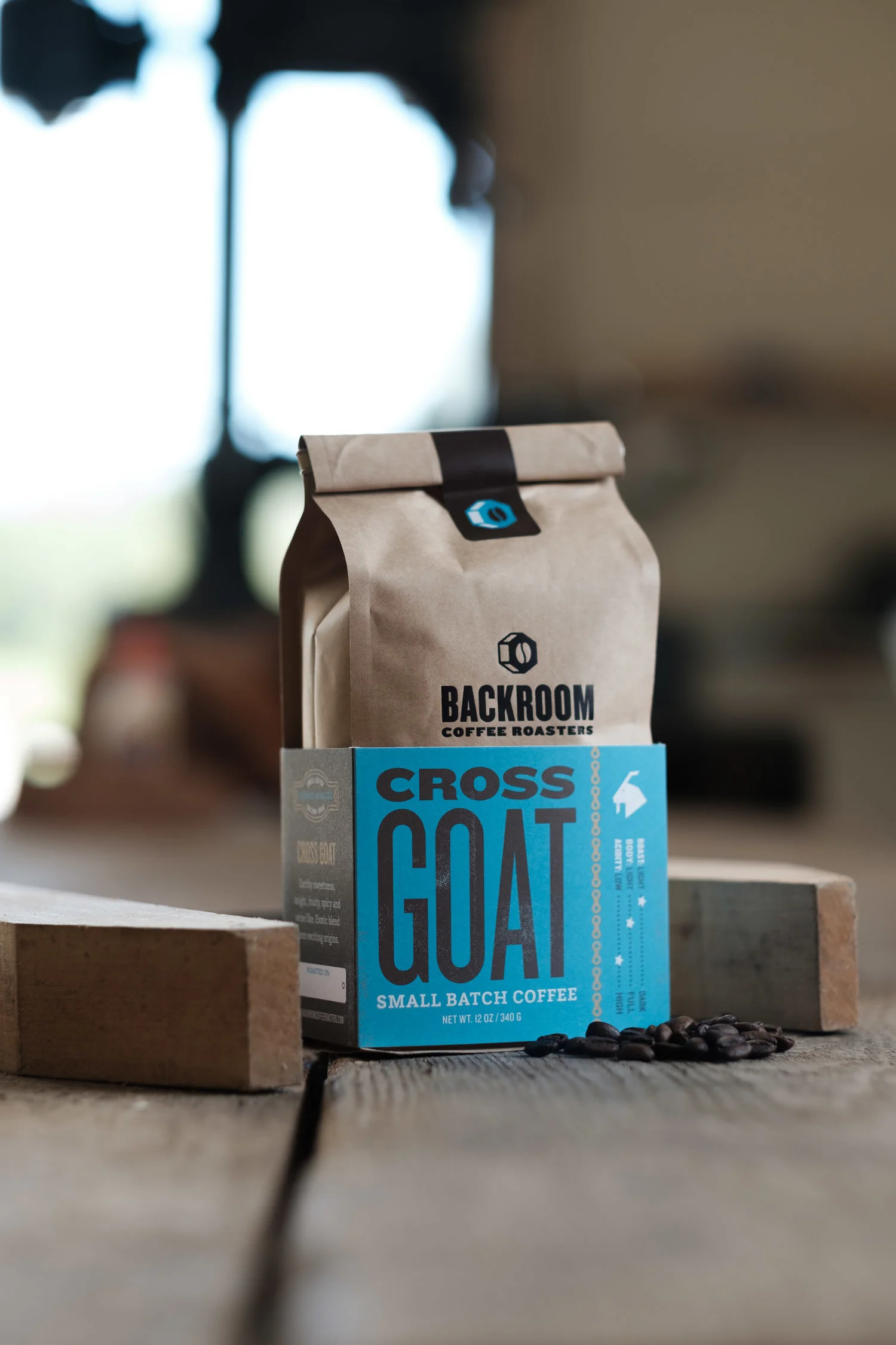 A brown paper bag of Backroom Coffee Roasters' Cross Goat small batch coffee on a wooden surface with some coffee beans scattered in front, against a blurred background.