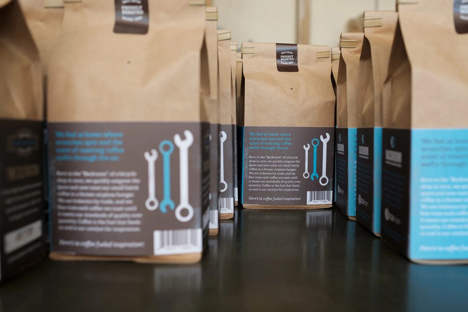 Multiple brown paper bags with black and blue labels on a black surface, used for packaging coffee.
