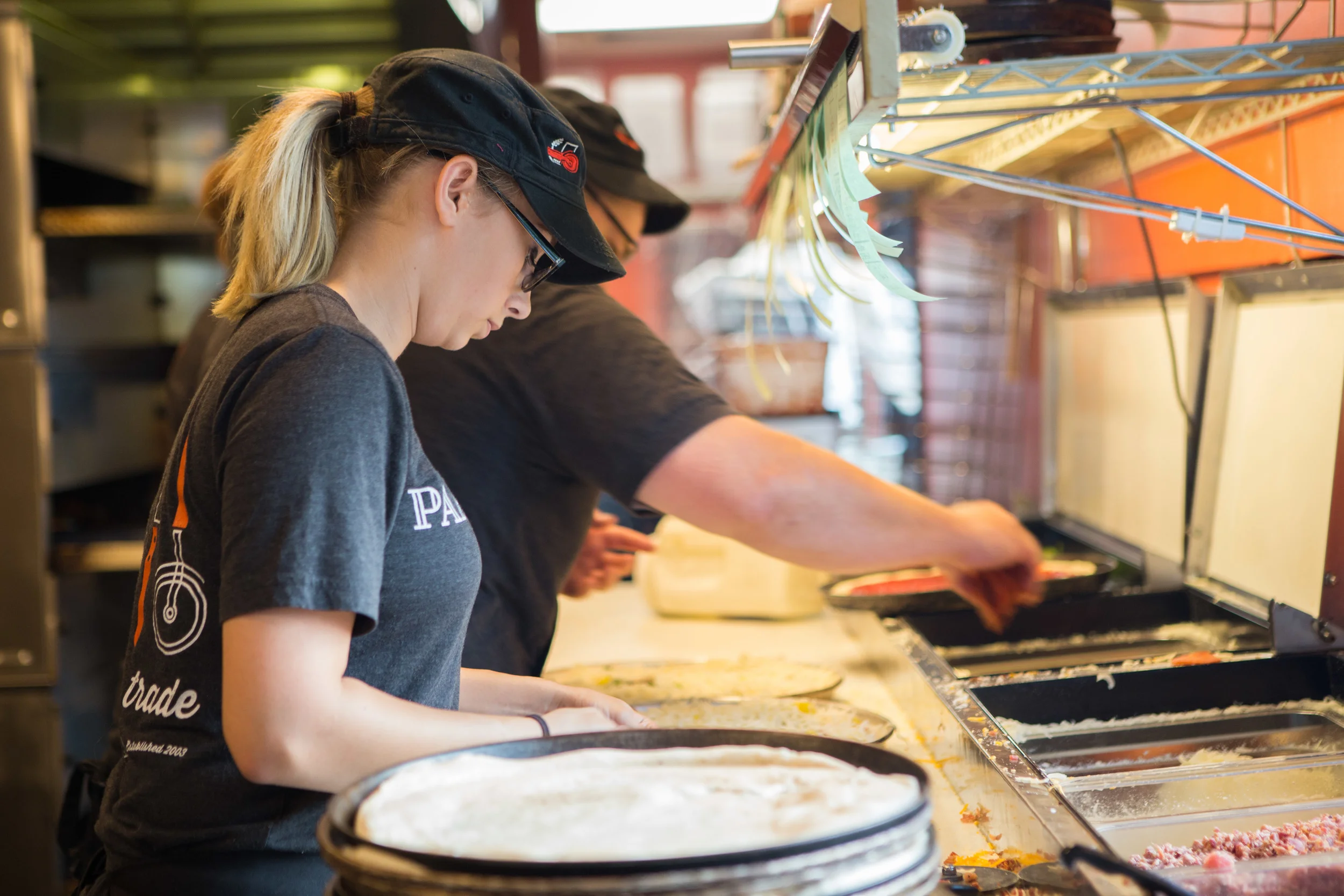 A young woman preparing pizzas at a pizza shop, wearing a black cap and T-shirt, with a focused expression, along with another worker in the background.