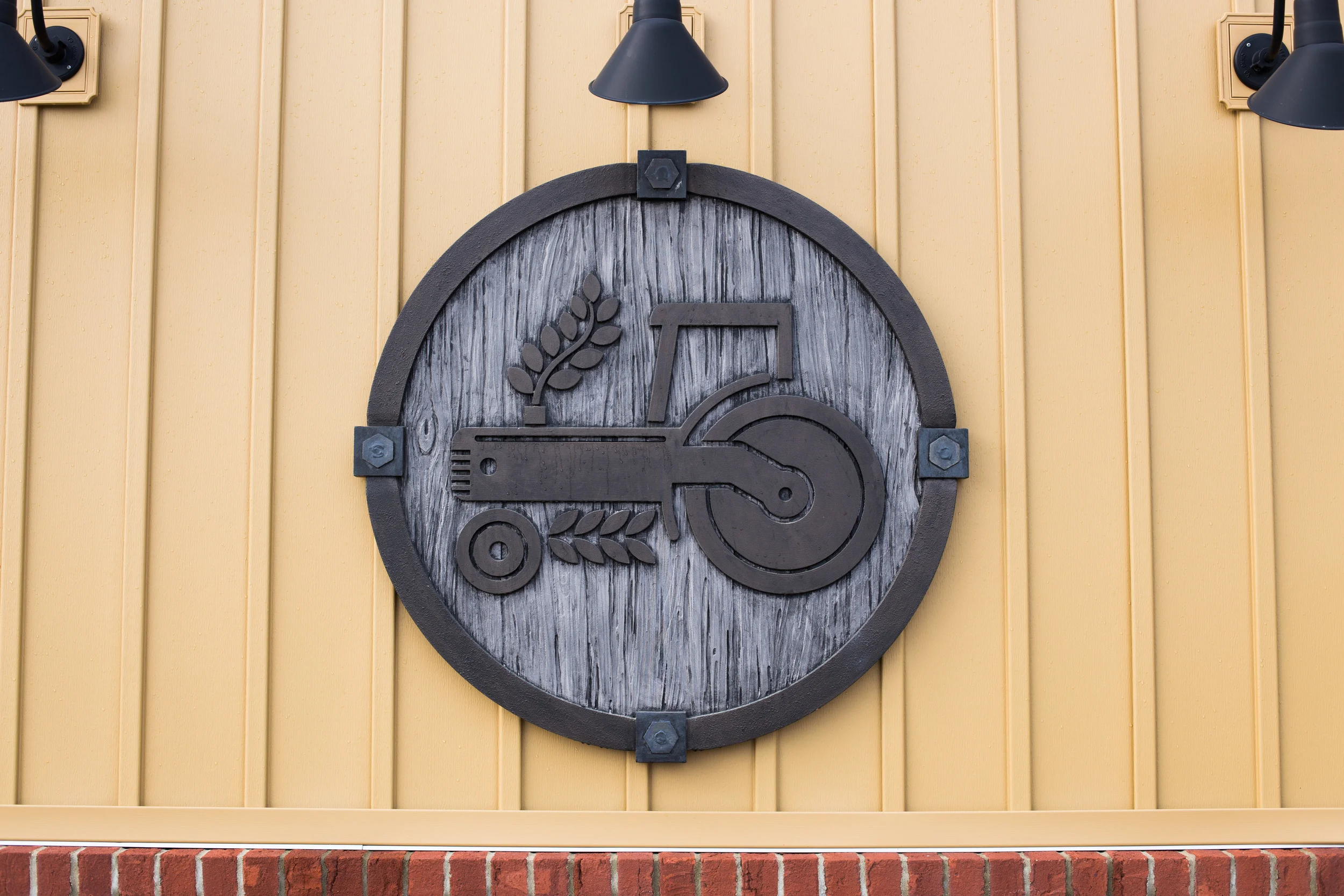 A decorative sign featuring a circular wooden background with a black tractor and wheat design mounted on a yellow wooden wall, illuminated by two black outdoor lights.