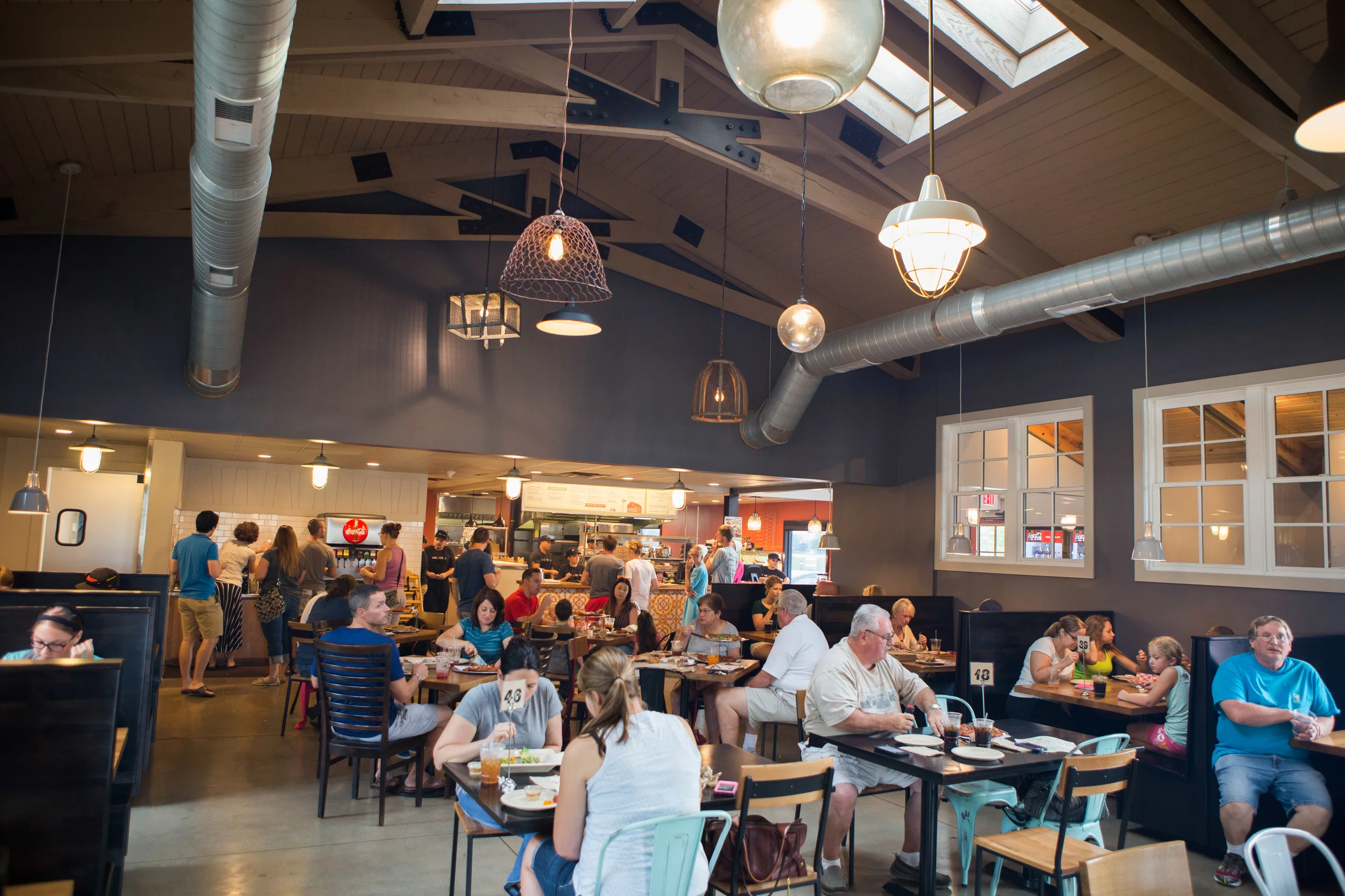 Interior of a casual restaurant with diners seated at tables and a busy open kitchen in the background. The ceiling features exposed wooden beams, hanging lights, and large air ducts.