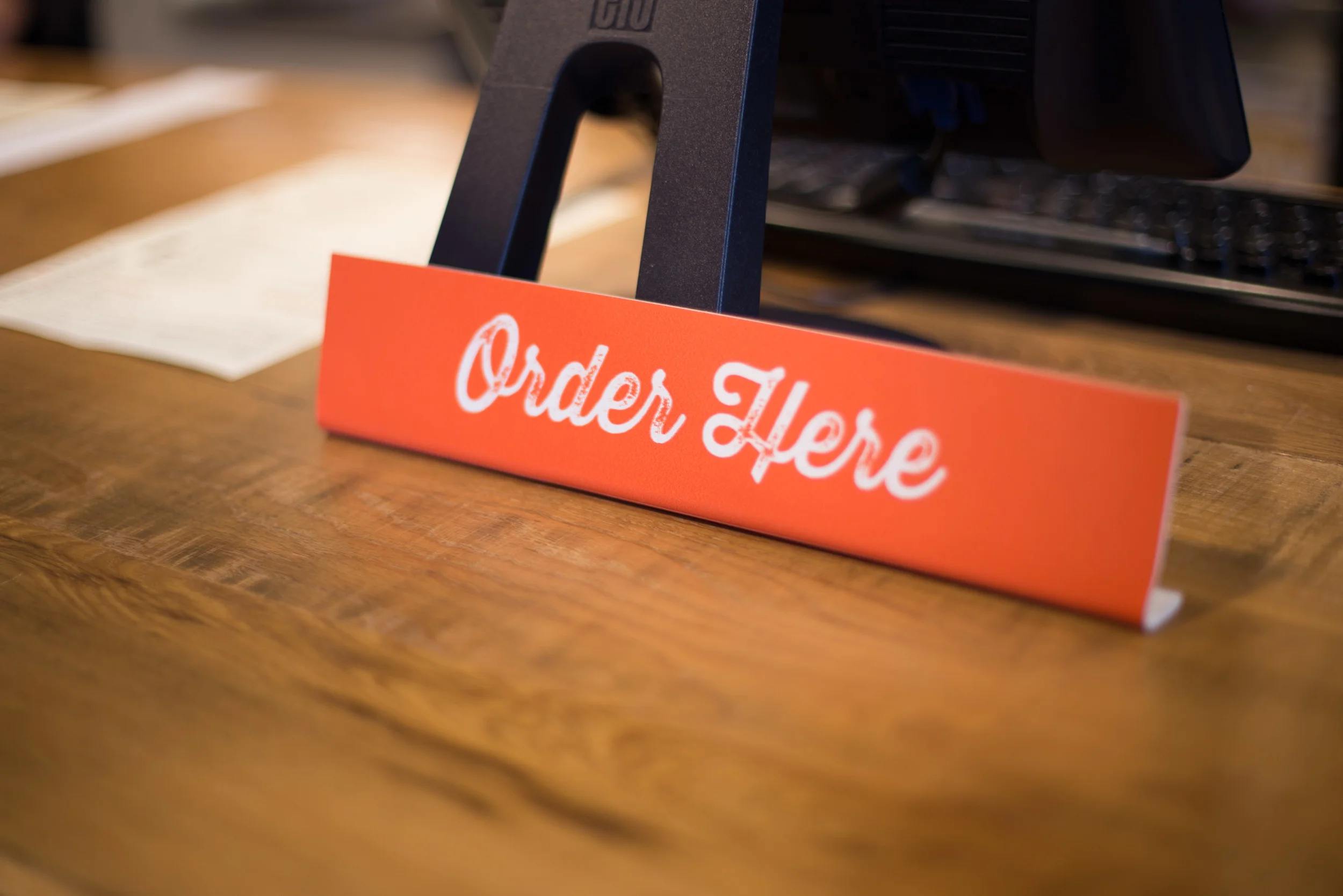 Red sign on a wooden counter with white cursive text that says "Order Here."