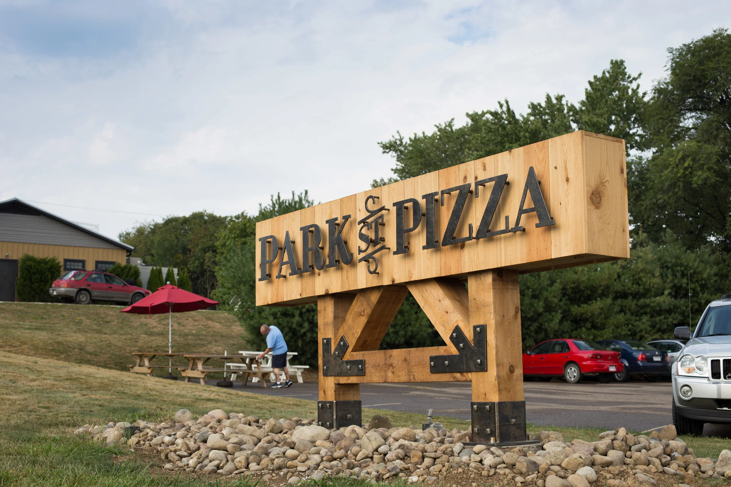 A large wooden sign for Park & Pizza with black letters, located on a grassy area with rocks, in front of a parking lot with cars, trees, and a building in the background.