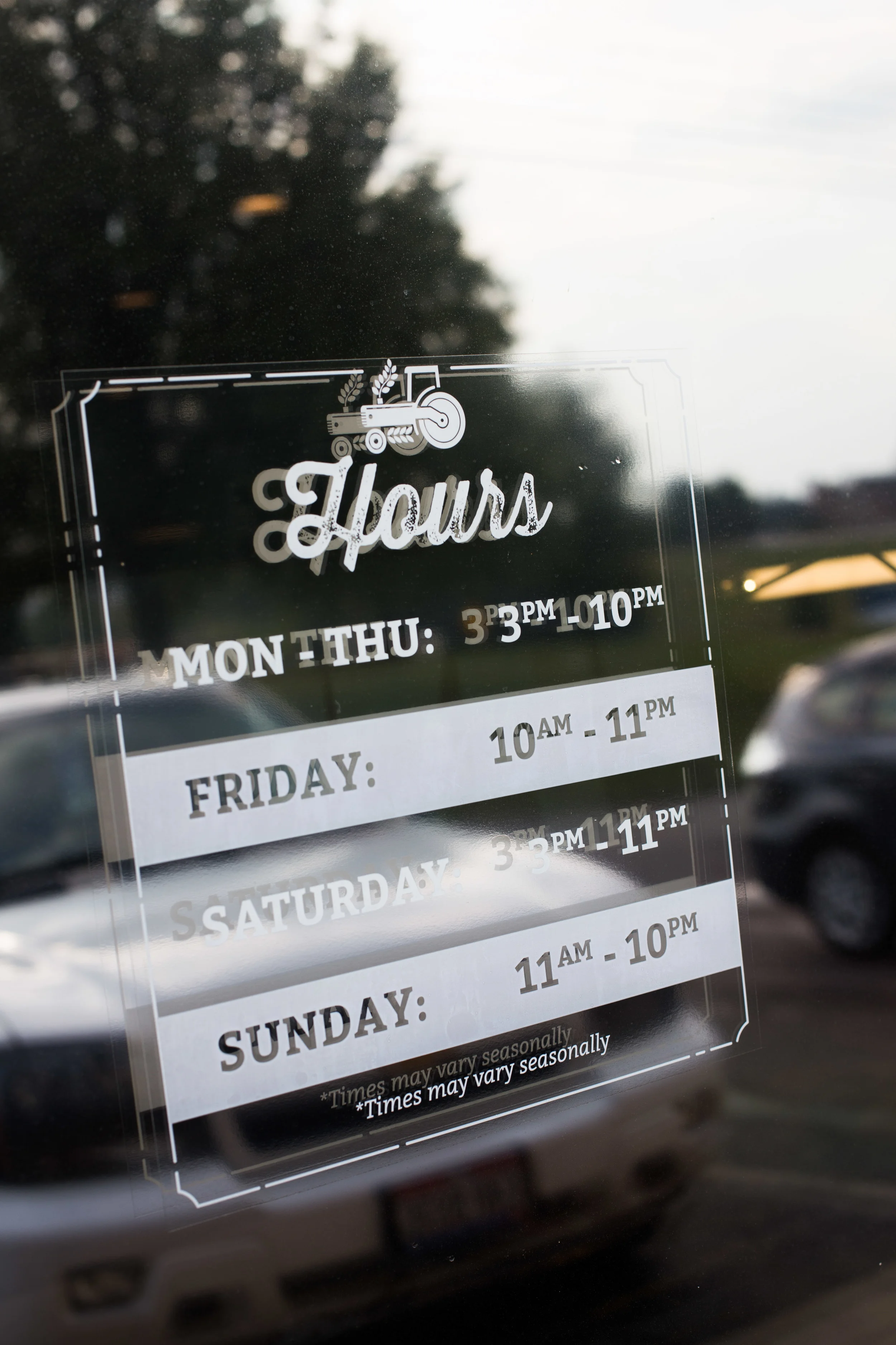 Sign on a glass window displaying business hours for Monday through Sunday, with details varying by day. The sign features a small tractor graphic at the top.