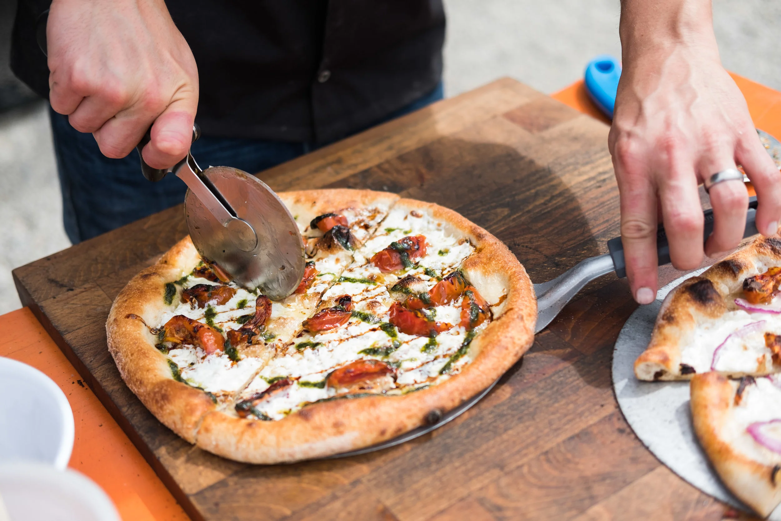 Person slicing a freshly baked pizza with tomato, basil, and mozzarella cheese on a wooden table.