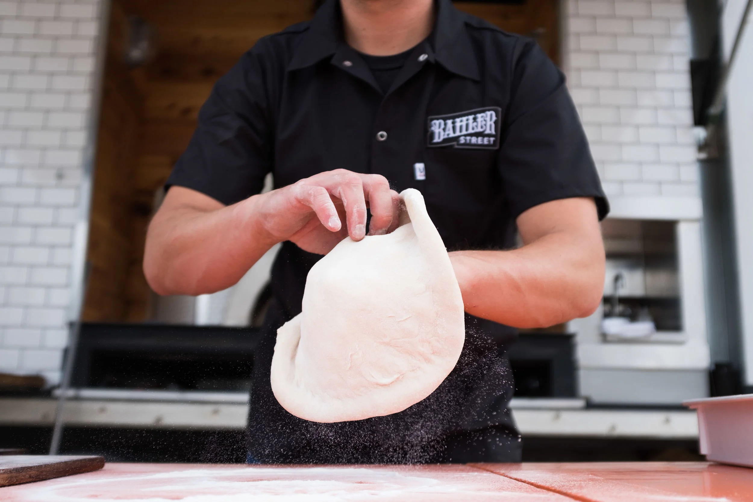 Person tossing pizza dough in the air in a kitchen, wearing a black shirt with a patch that says 'Batter Street'.