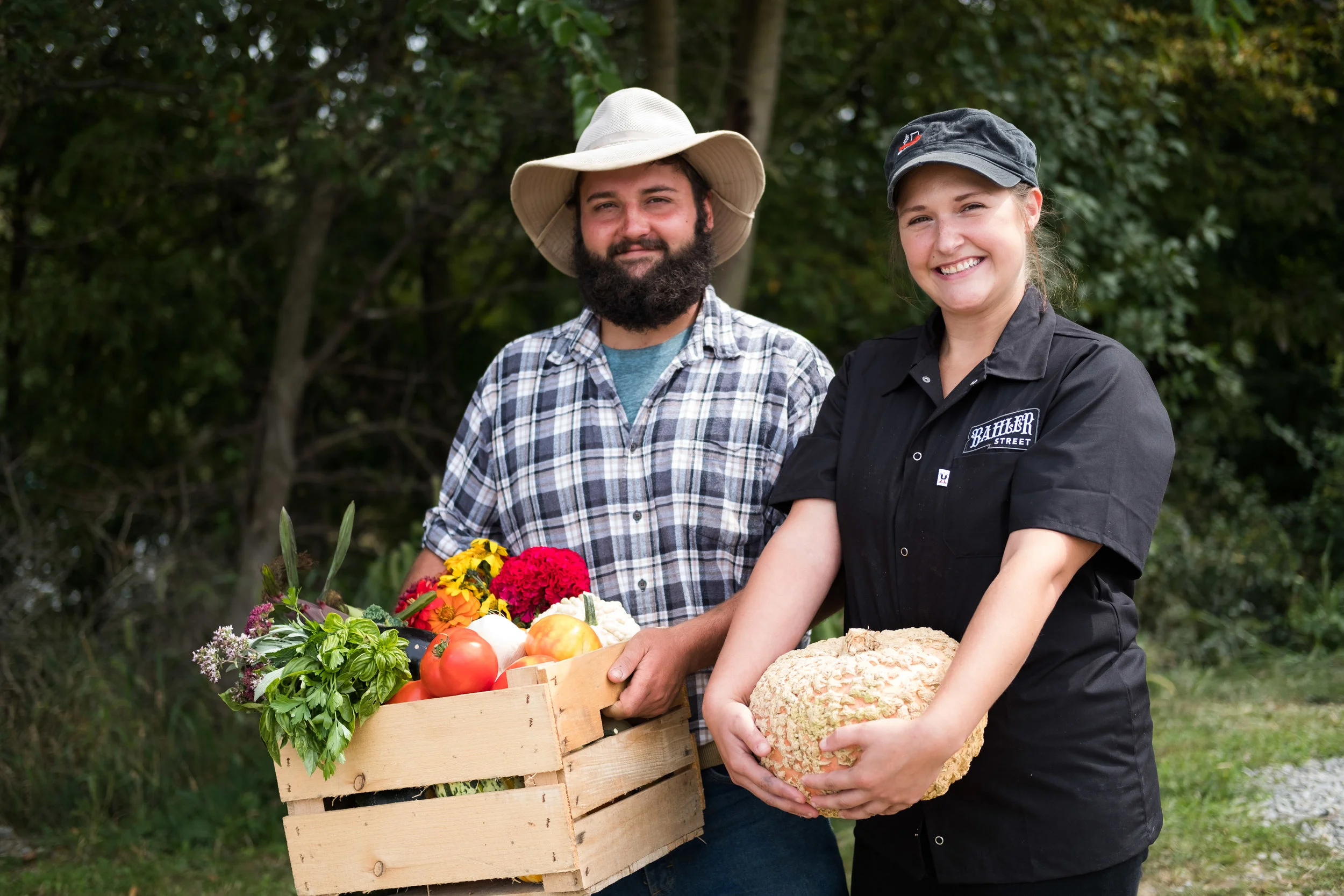 Smiling man with a beard wearing a plaid shirt and a wide-brimmed hat stands next to a smiling woman with a black uniform and cap, holding a large vegetable, outdoors with trees in the background. The man holds a wooden crate filled with fresh vegeta