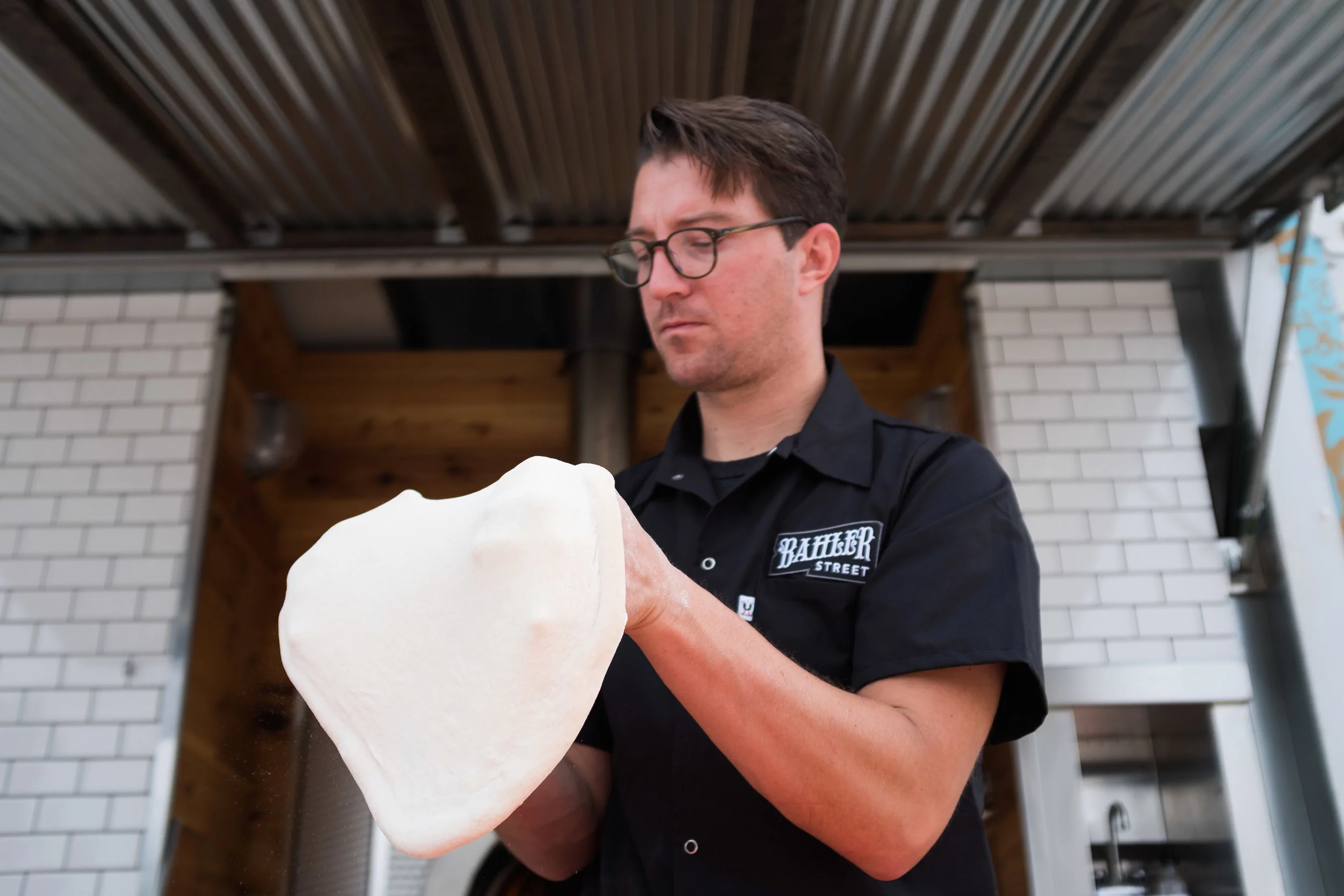 Man with glasses and a black shirt holding a large piece of pizza dough