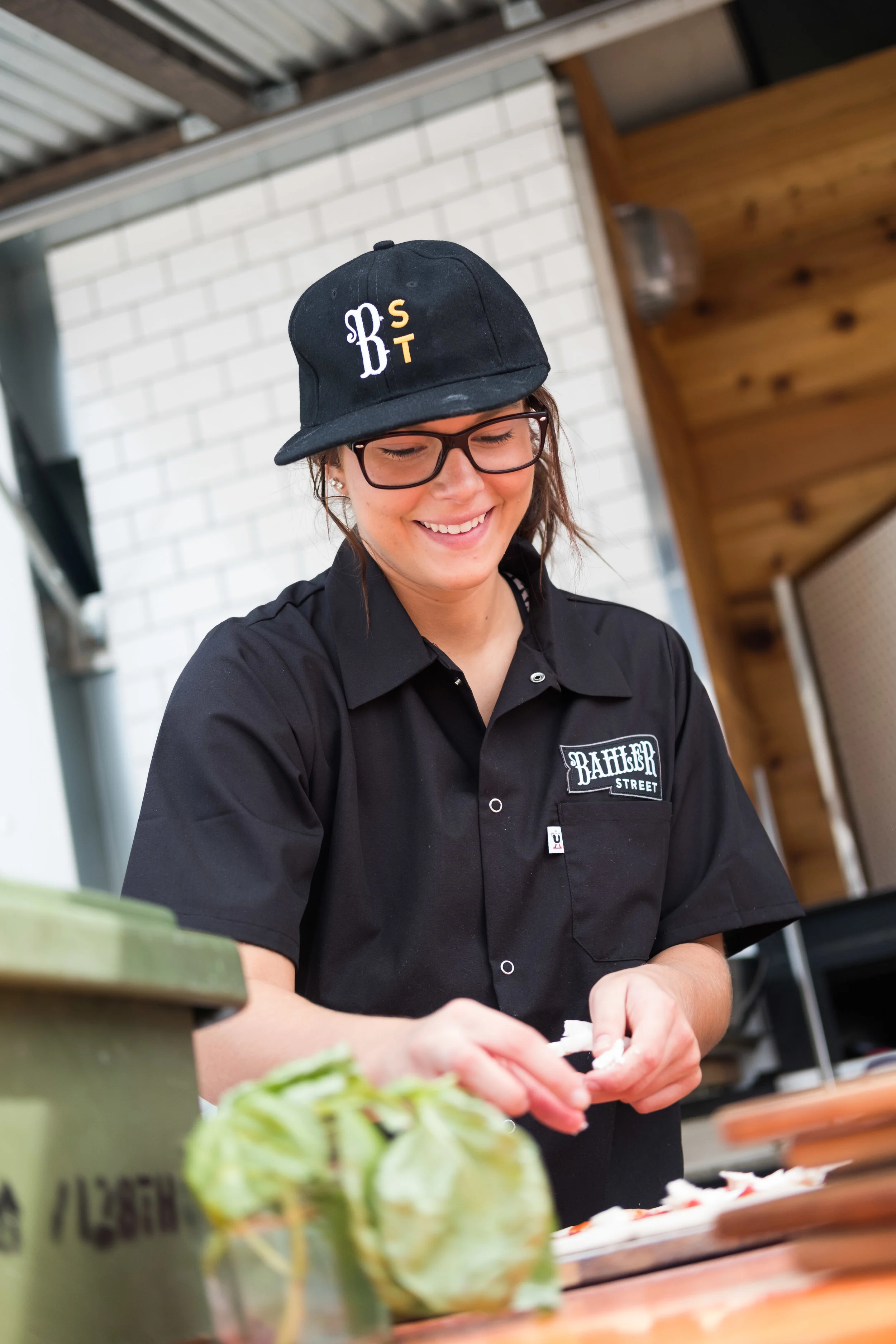 A smiling woman wearing glasses, a black baseball cap, and a black shirt with "RAIDER STREET" logo, preparing food in a rustic kitchen with white brick wall and wooden accents.
