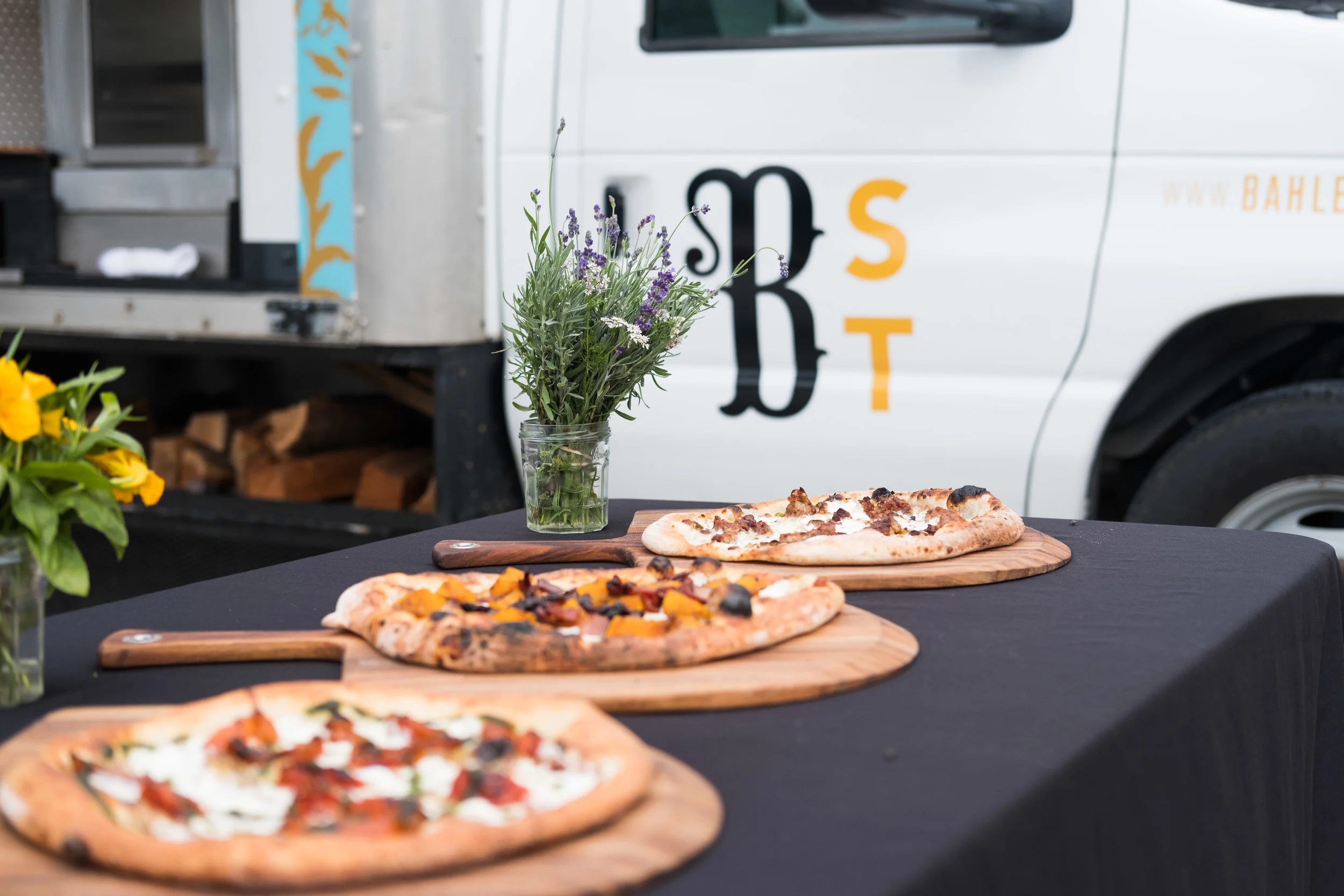 Three pizzas on wooden boards on a black tablecloth, with a small vase of lavender and other flowers nearby, set in front of a food truck with partially visible logo and a wheel.