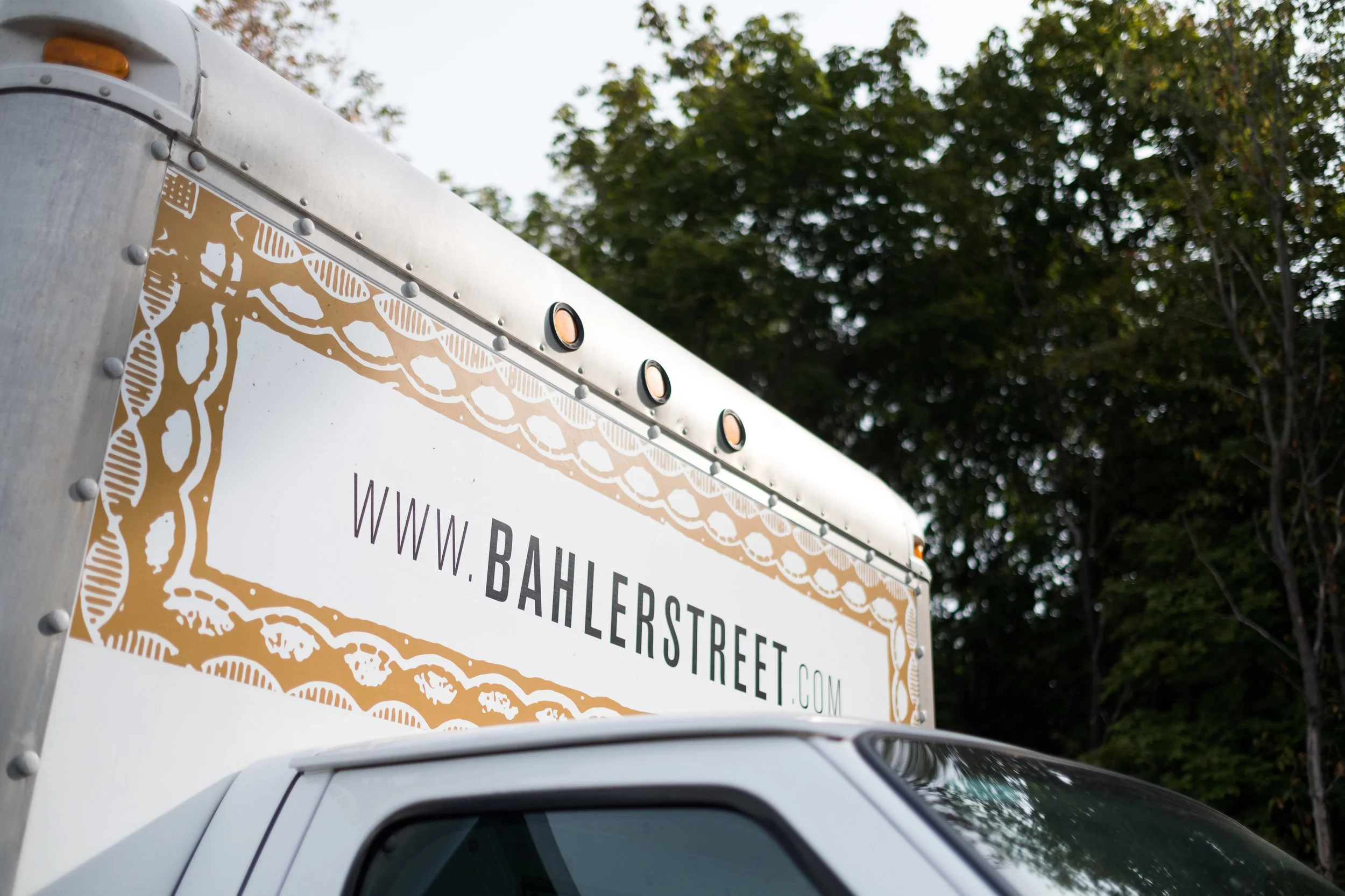 Close-up of a delivery truck with a sign displaying the website www.bahlerstreet.com, part of the truck's side and front window, with a background of green trees.