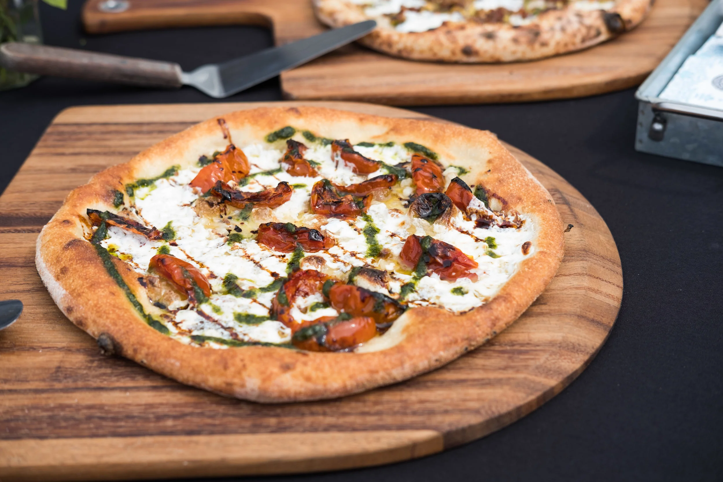 Close-up of a wood-fired pizza topped with cherry tomatoes, mozzarella cheese, and basil sauce on a wooden pizza board.