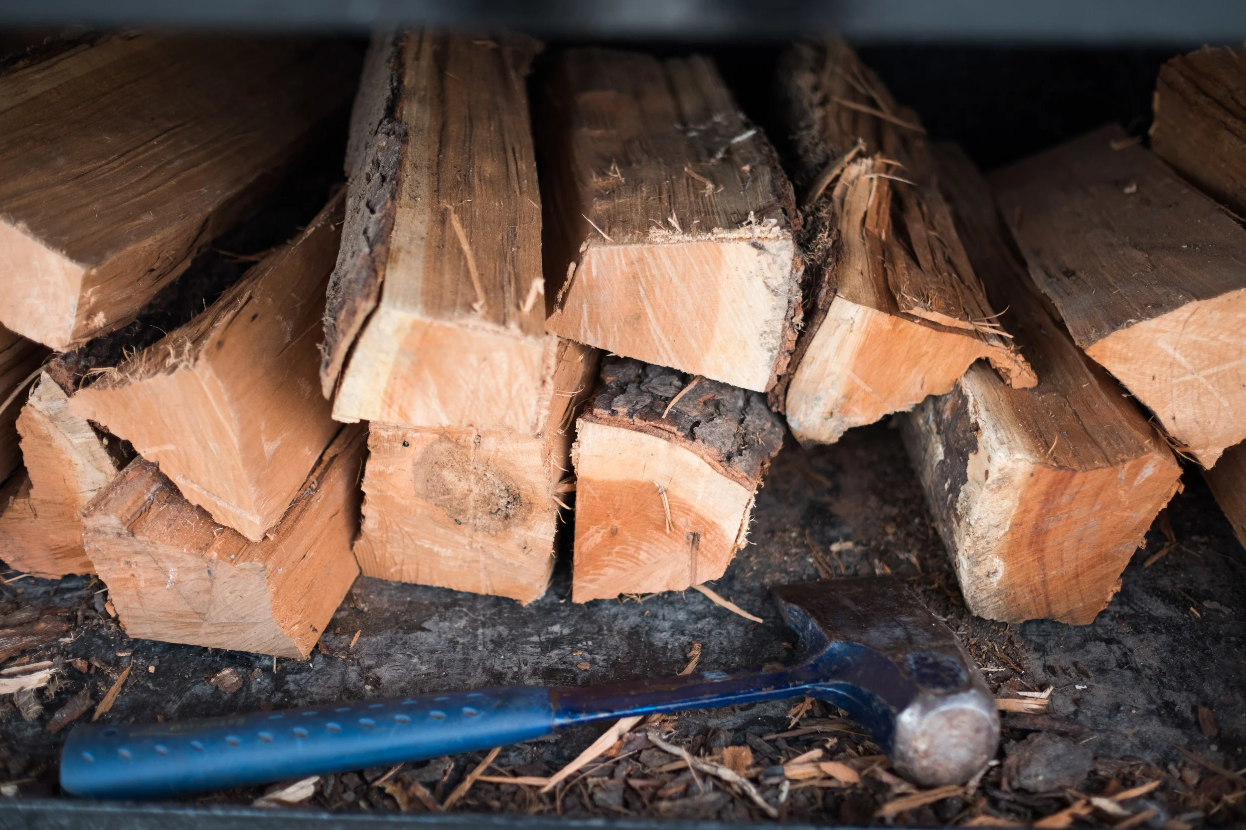 Stacked chopped firewood logs with a hammer lying on the ground.