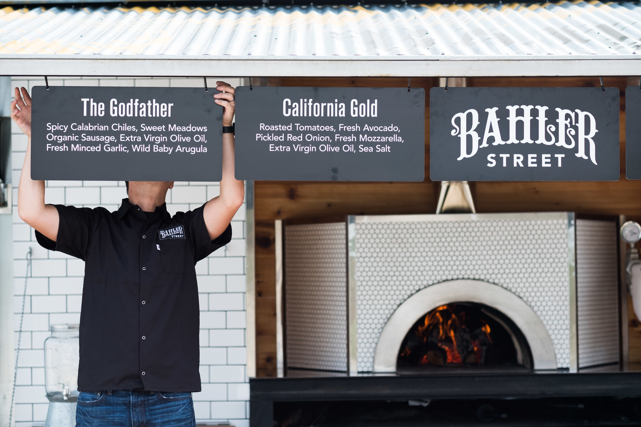 A person wearing a black shirt with a 'Bailbreaker Street' logo holds up two chalkboard signs at a pizzeria. The signs list menu items: 'The Godfather' and 'California Gold.' A wood-fired pizza oven is visible beneath the signs.