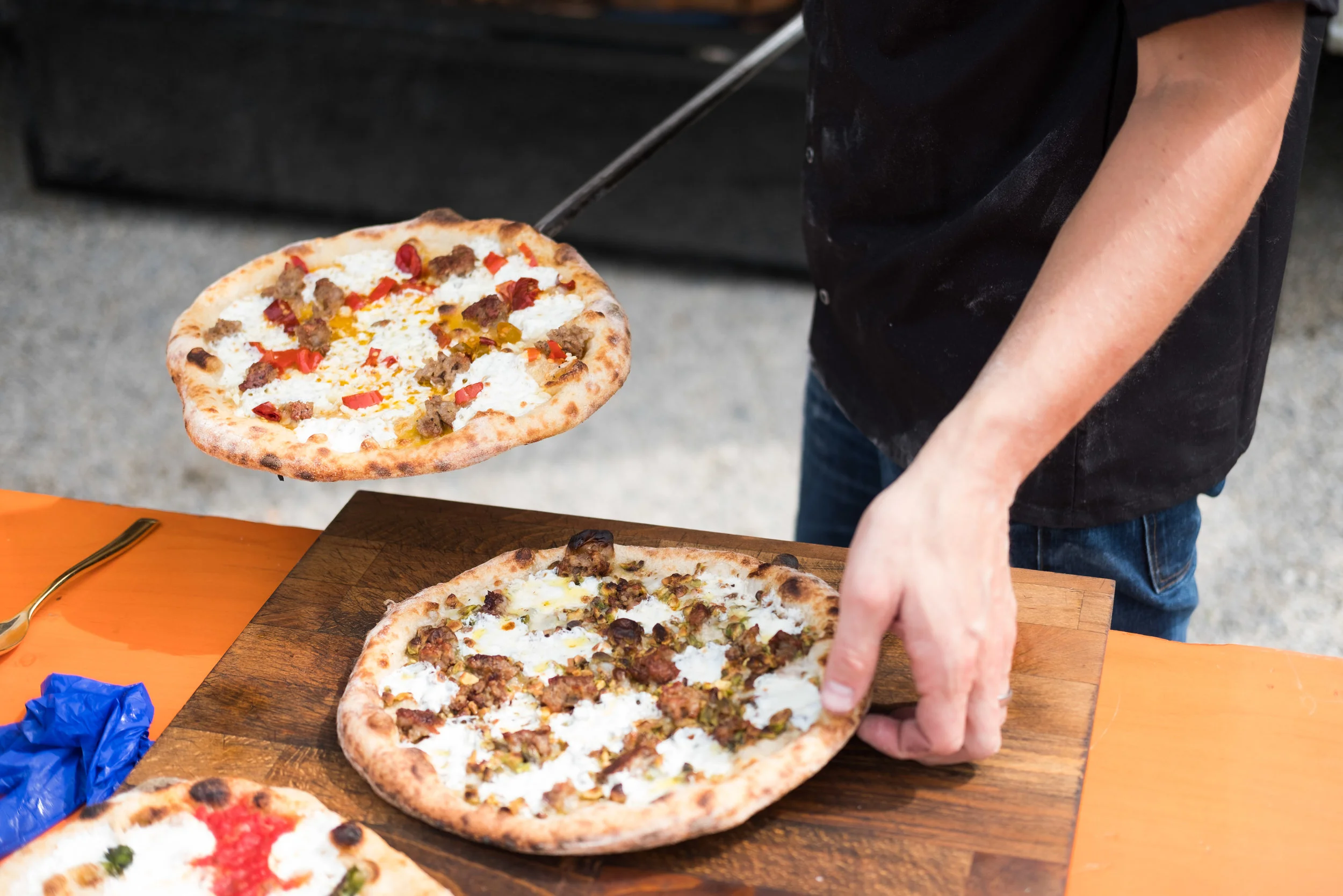 Person removing a pizza from a wooden board at an outdoor setting with other pizzas and a table.