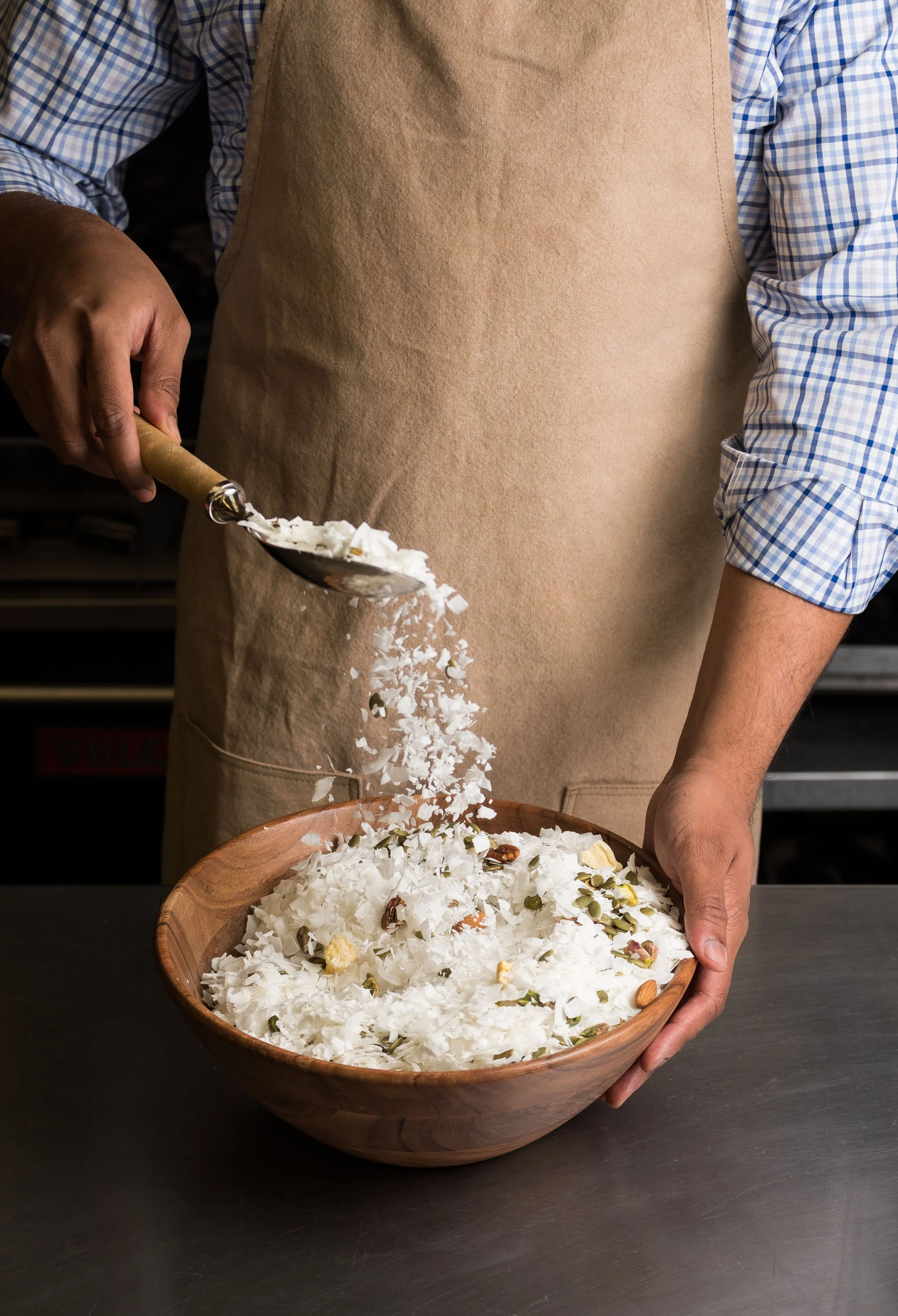A person wearing a checkered shirt and beige apron sprinkling shredded coconut and nuts from a scoop into a wooden bowl.