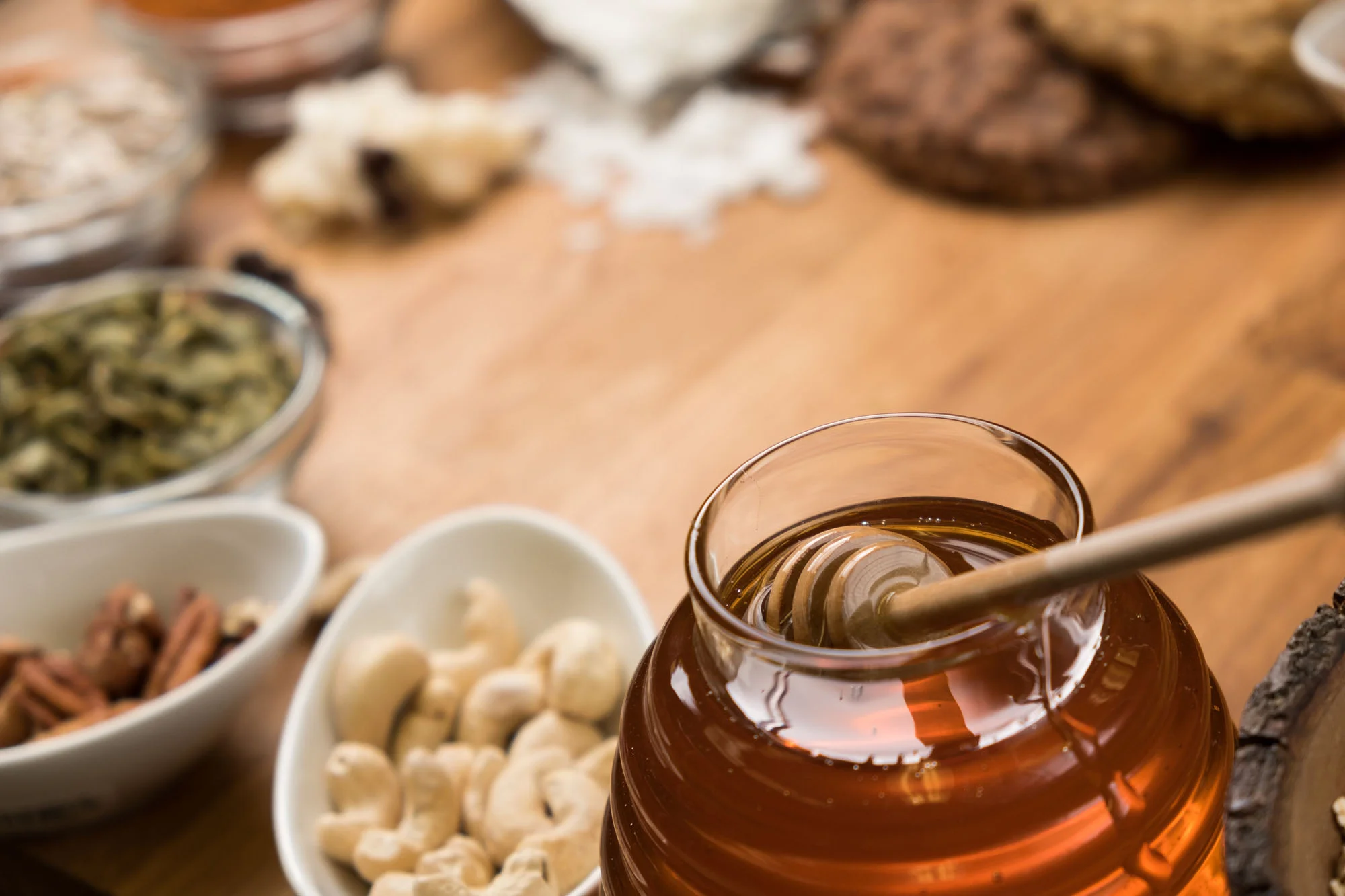 A jar of honey with a honey dipper in it, surrounded by bowls of various nuts and dried fruits on a wooden surface.