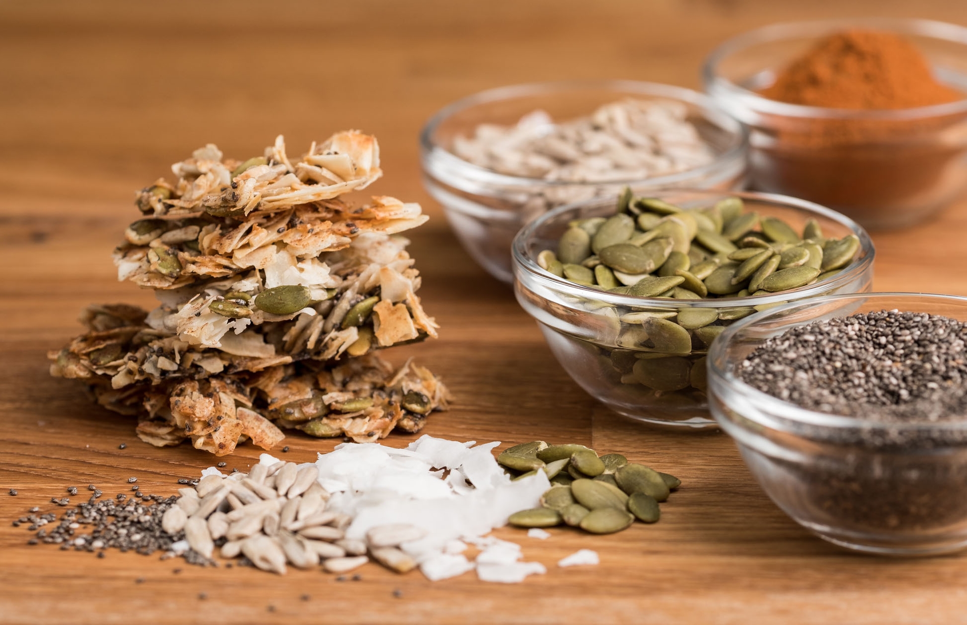 Granola bars and various seeds and nuts in glass bowls on wooden surface.