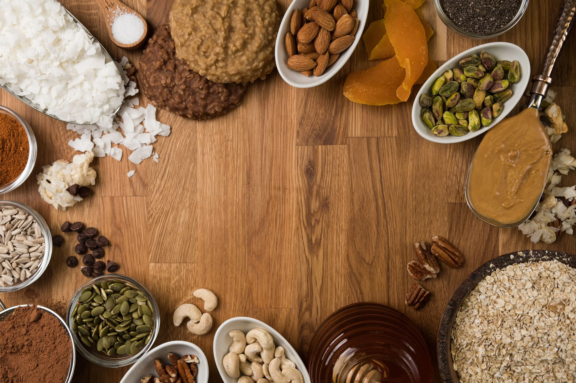 Assorted ingredients on a wooden table, including coconut flakes, crushed cookies, spices, nuts, dried fruits, pumpkin seeds, sunflower seeds, cacao nibs, peanut butter, dried apricots, pecans, cashews, and rolled oats.