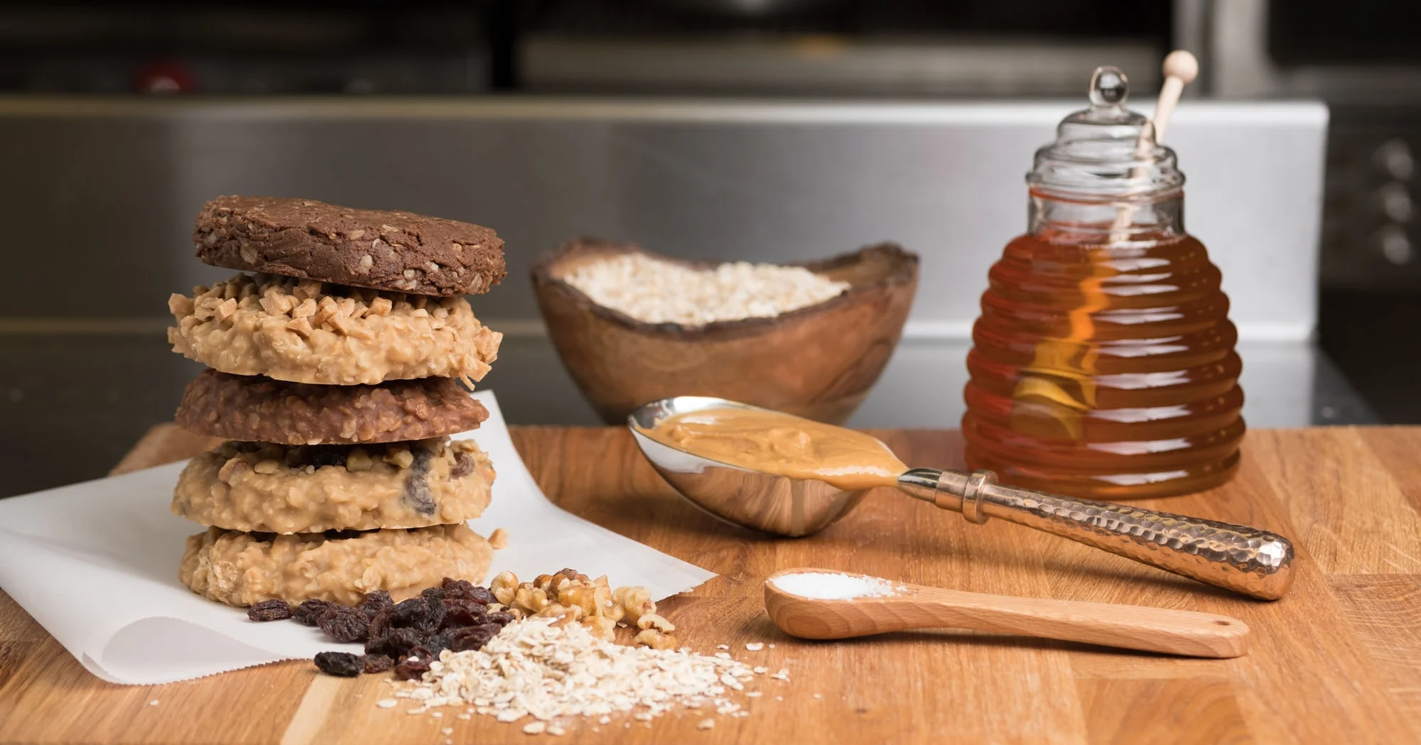 Stacked cookies on parchment paper, a bowl of coconut flakes, dried raisins, a spoonful of peanut butter, a honey jar, and a small wooden scoop with flour on a wooden surface.