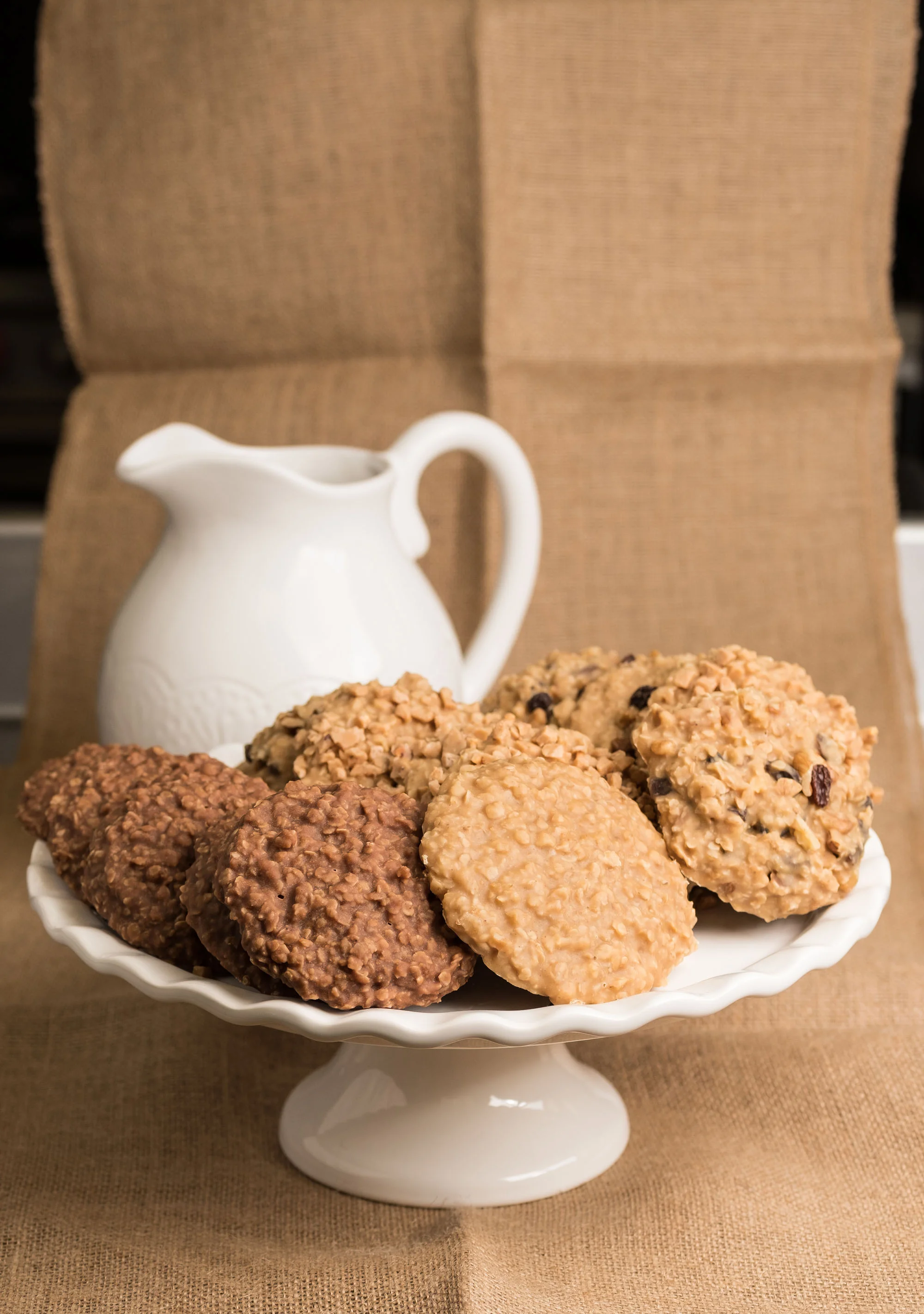 A white pedestal dish filled with assorted oatmeal cookies, placed in front of a white ceramic pitcher on a brown fabric-covered surface.