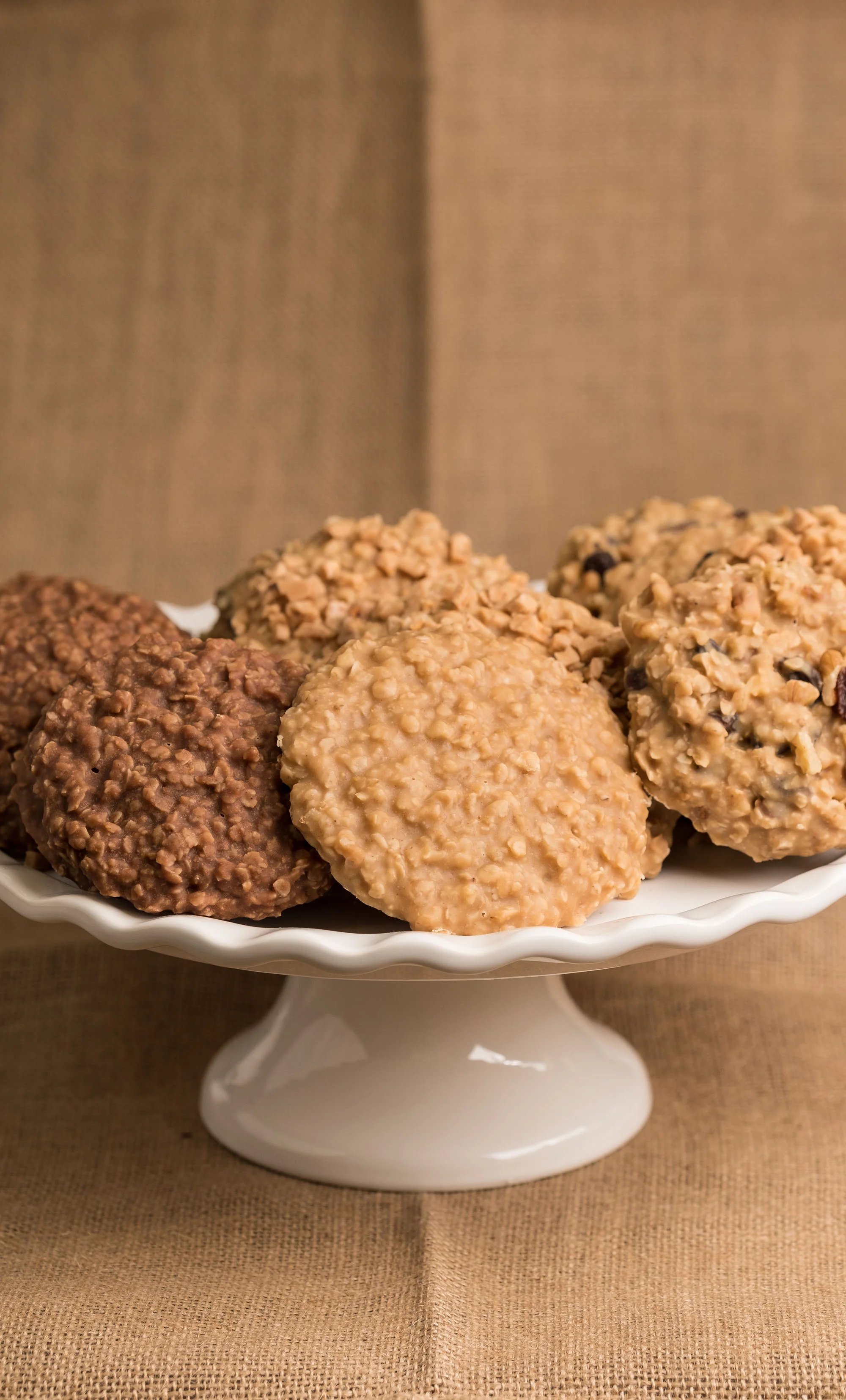 A white cake stand with assorted cookies, including oatmeal and chocolate chip, on a brown fabric surface.