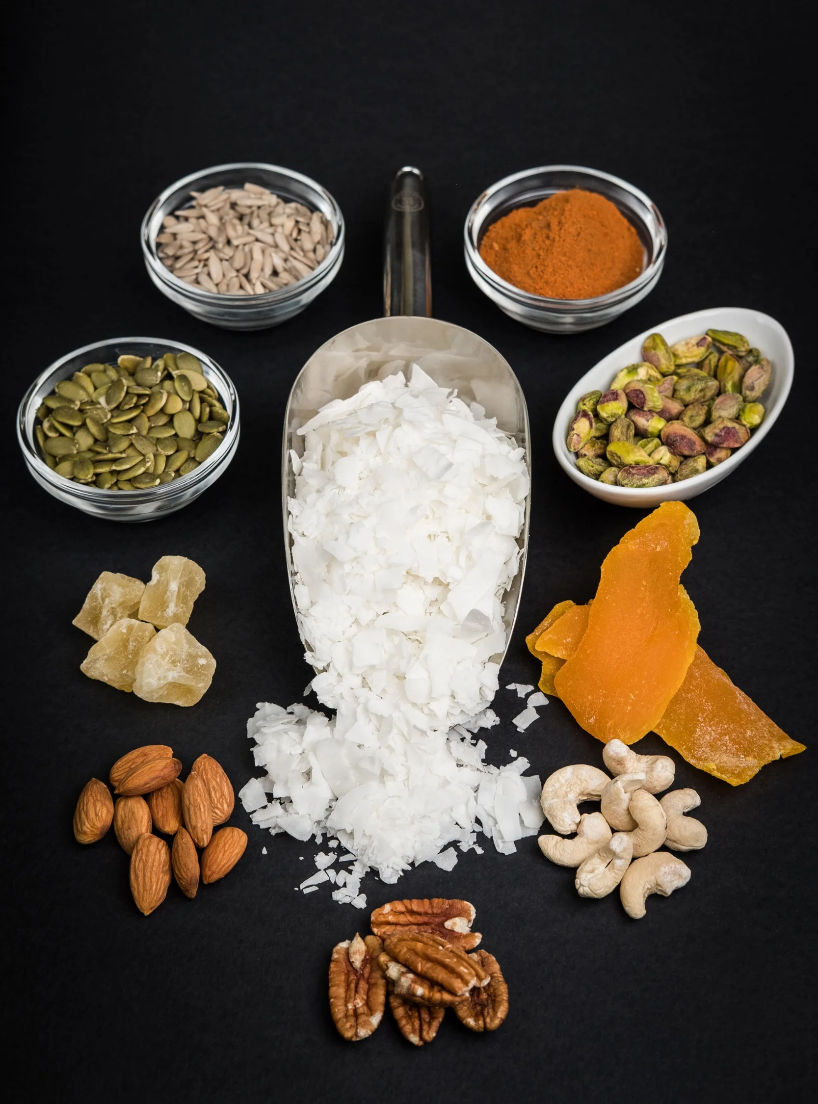 Assorted ingredients including sunflower seeds, turmeric powder, pumpkin seeds, coconut flakes, dried ginger, sugar cubes, almonds, cashews, pecans, dried fruit, and other nuts arranged on a black surface.