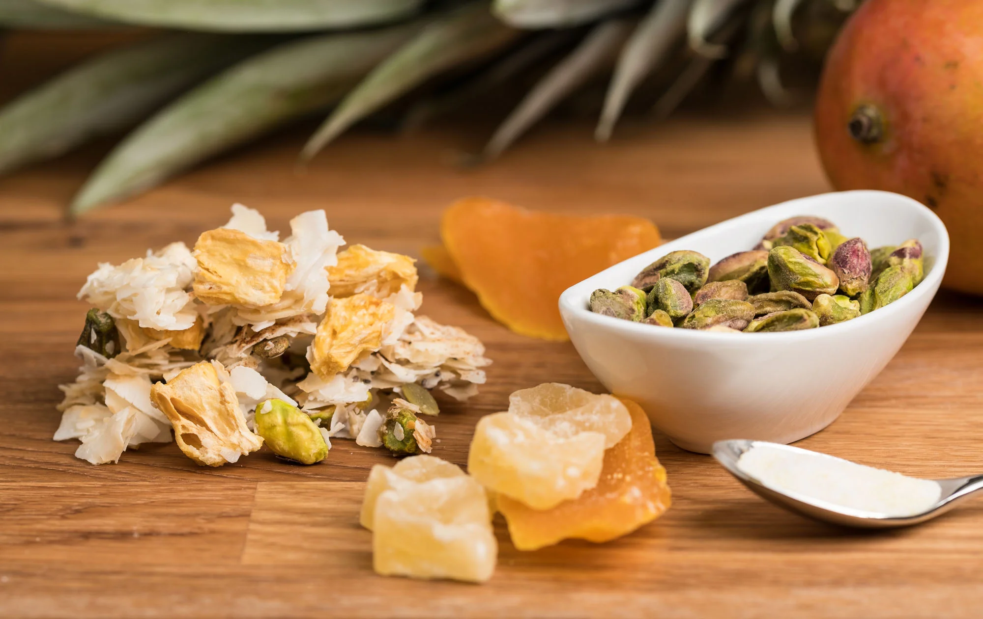A selection of dried fruit and nuts on a wooden surface, including pineapple chunks, dried papaya, candied ginger, and pistachios in a small white bowl, with a pineapple in the background.