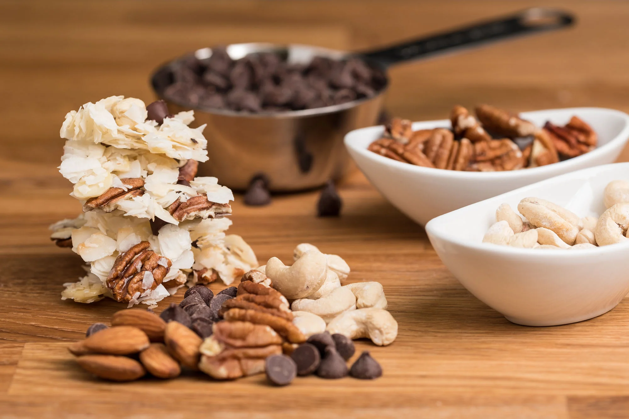 Various nuts, chocolate chips, and white chocolate bark on a wooden surface with bowls of nuts and chocolate in the background.