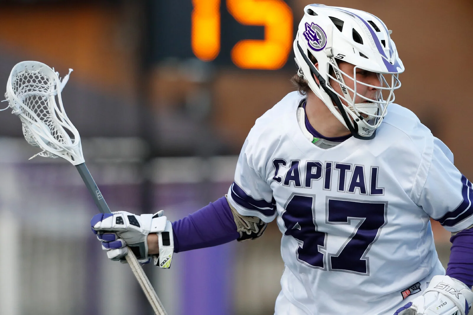 Lacrosse player wearing a white jersey with purple text that reads 'CAPITAL' and the number 42, holding a lacrosse stick with a white net, wearing a helmet with faceguard, gloves, and purple sleeves, on a field during a game.