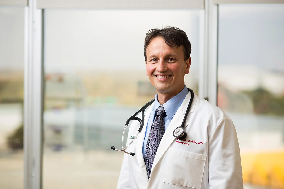 A smiling male doctor in a white lab coat with a stethoscope around his neck standing in front of a window