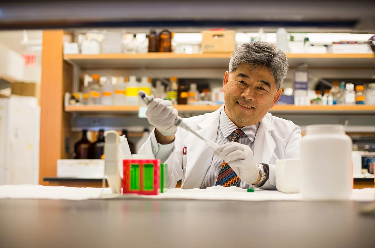 A male scientist in a white lab coat and gloves working with a pipette in a laboratory with various bottles and equipment on shelves behind him.
