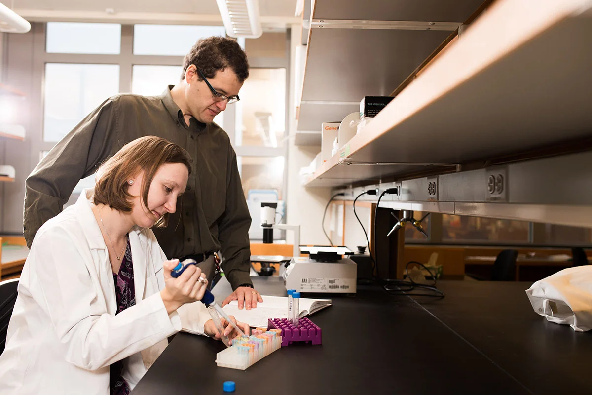 Two scientists working in a laboratory, one woman using a pipette and a man observing, with lab equipment and test tubes on the table.