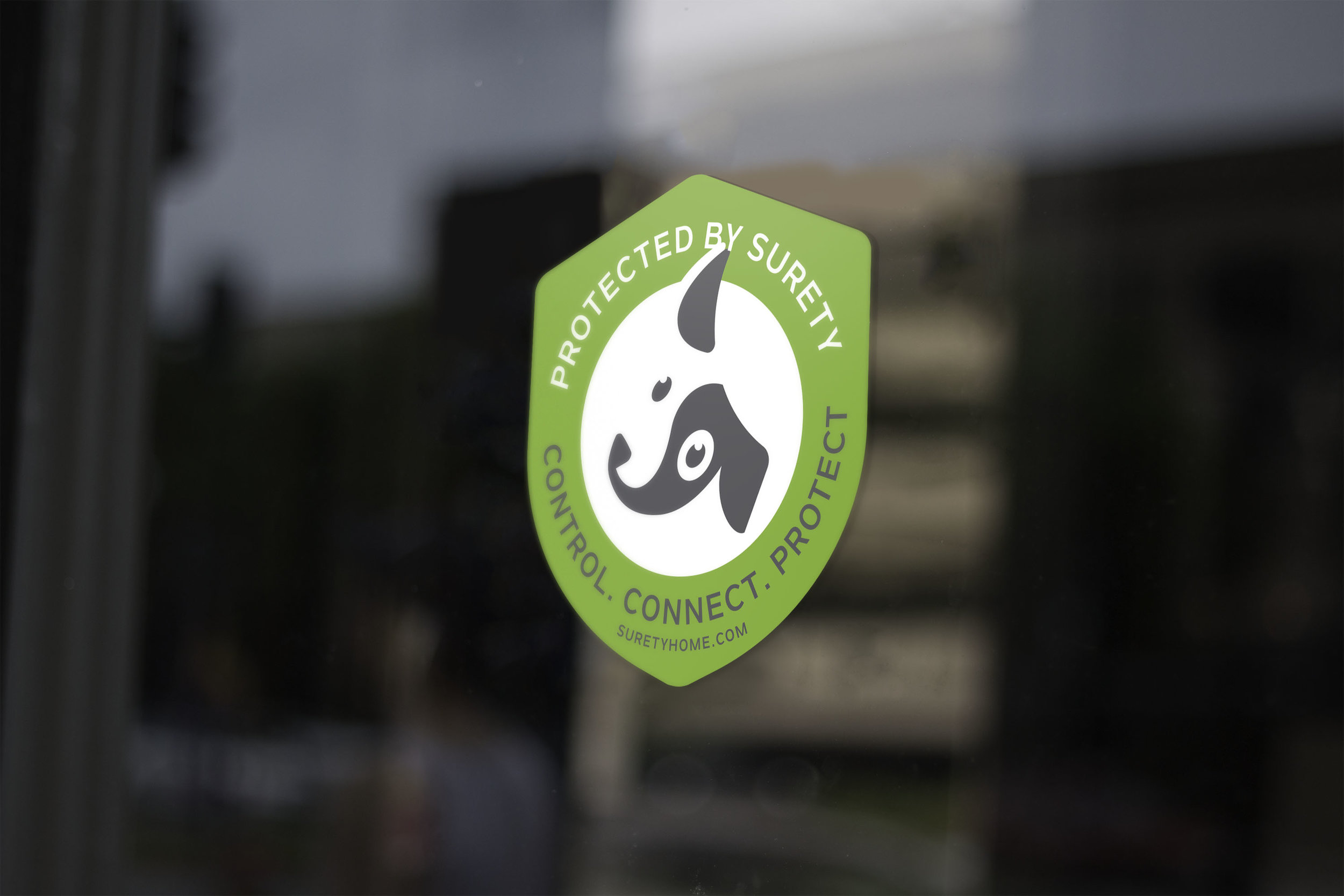 Green and white security sticker on a glass door with a dog icon and the words "Protected by Surety," "Control. Connect. Protect," and the website suretyhome.com.